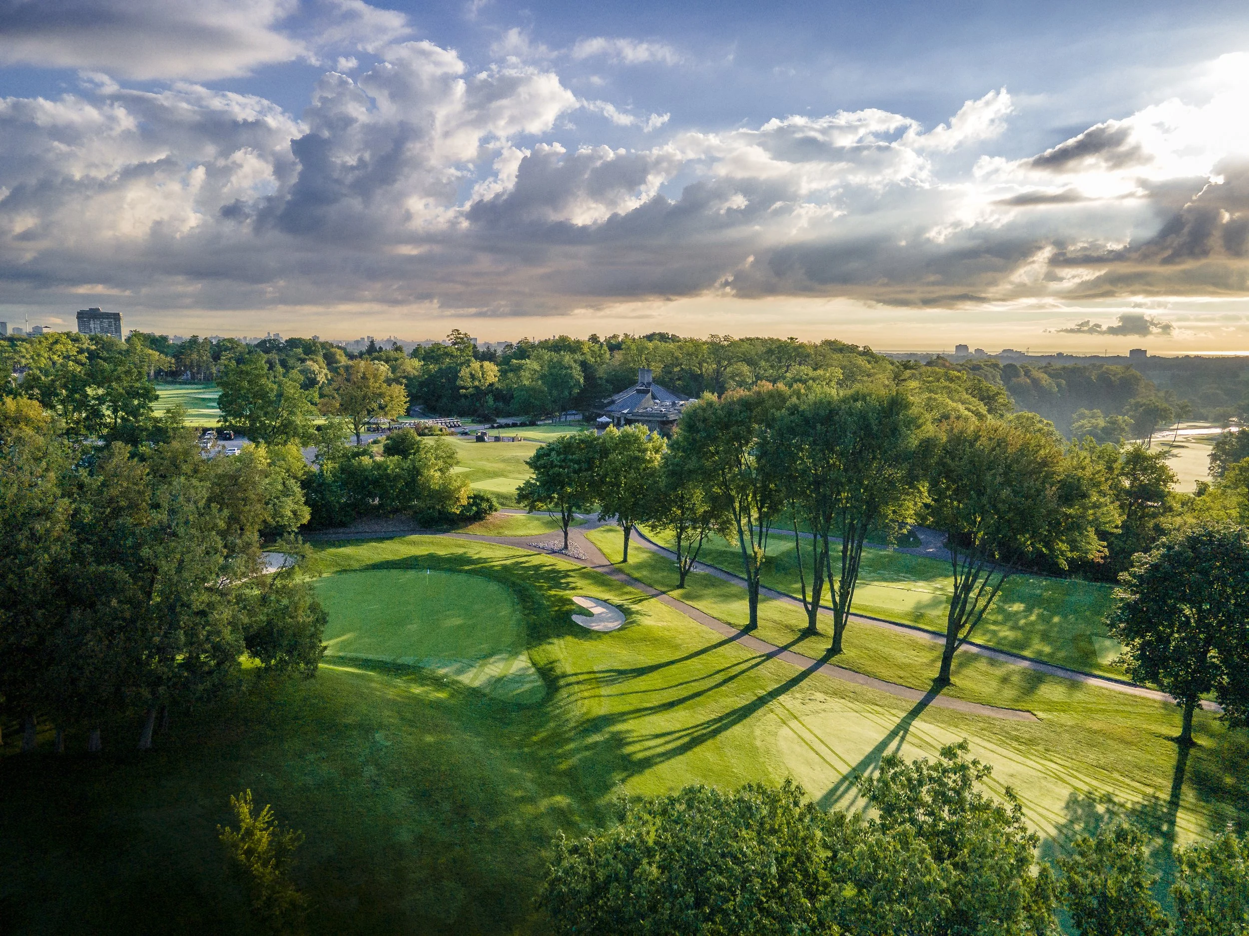 A scenic view of a golf course surrounded by trees, with a small building and parking lot, under a sky filled with clouds and sunlight.