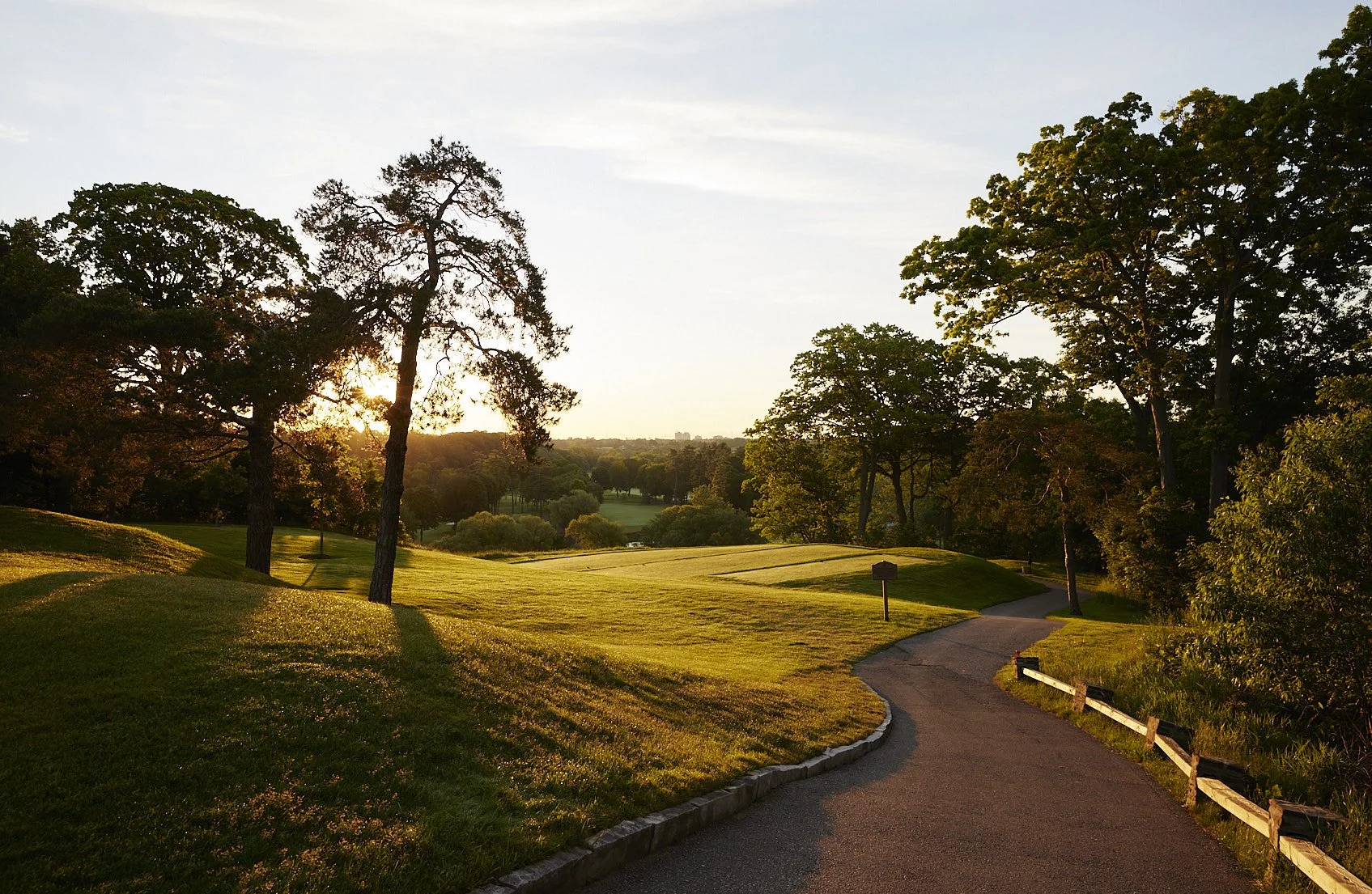 A peaceful golf hole at sunset with a winding path, grassy slopes, and tall trees casting long shadows.