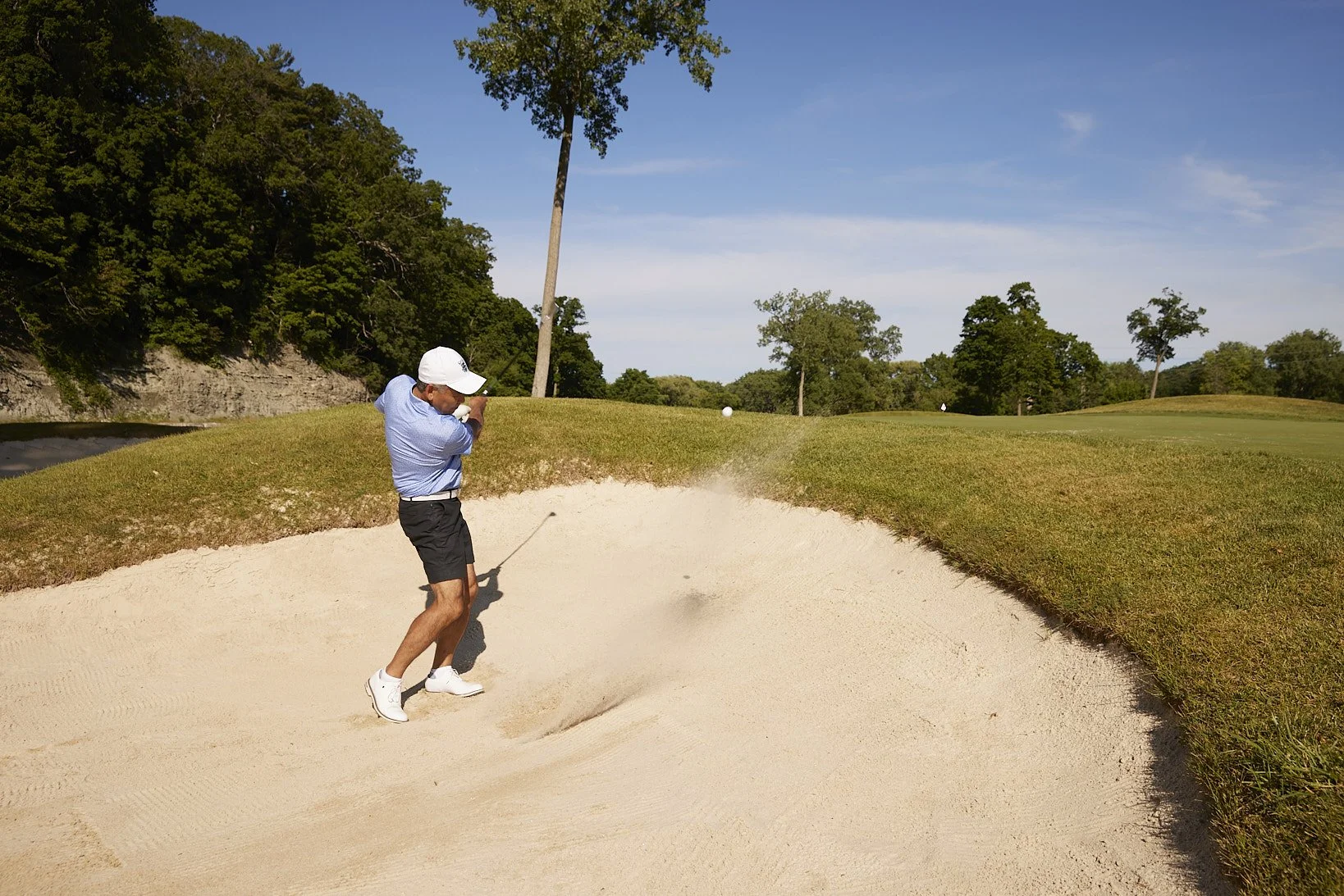 A man playing golf hitting a ball out of a sand bunker on a golf course during the day.