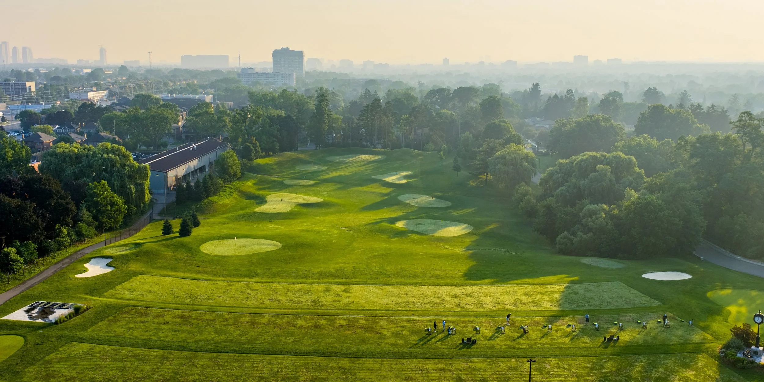 Aerial view of a driving range with multiple sand traps, surrounded by trees, with a city skyline in the distance and a hazy sky.