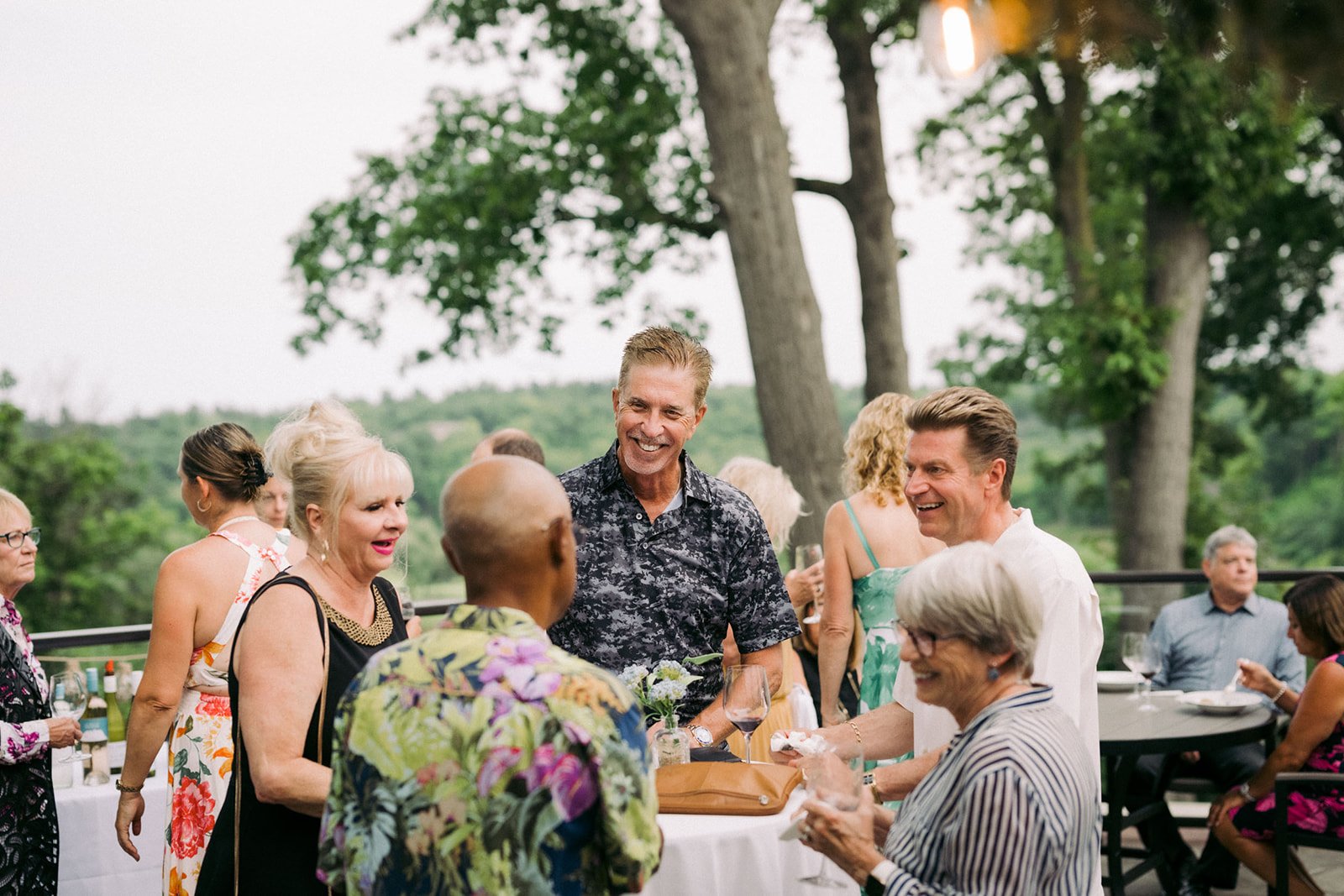 A group of people smiling and socializing at an outdoor gathering, with trees and greenery in the background, during daytime.