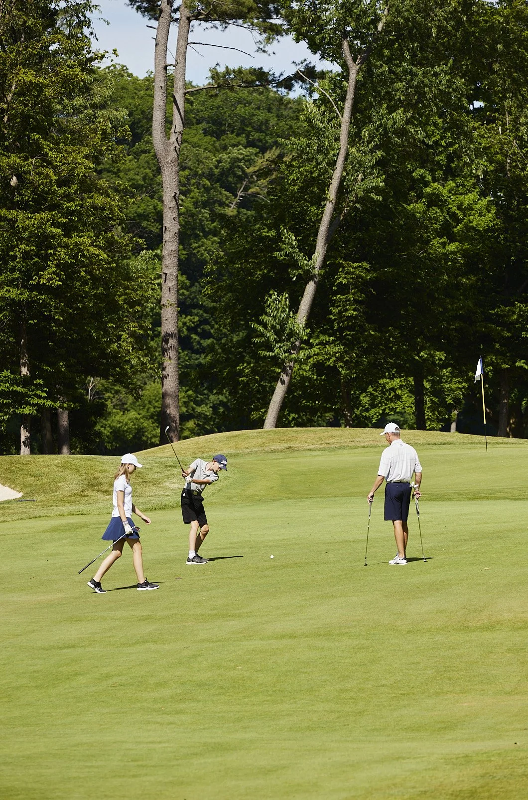 Three children playing mini-golf on a sunny day, surrounded by lush green trees.