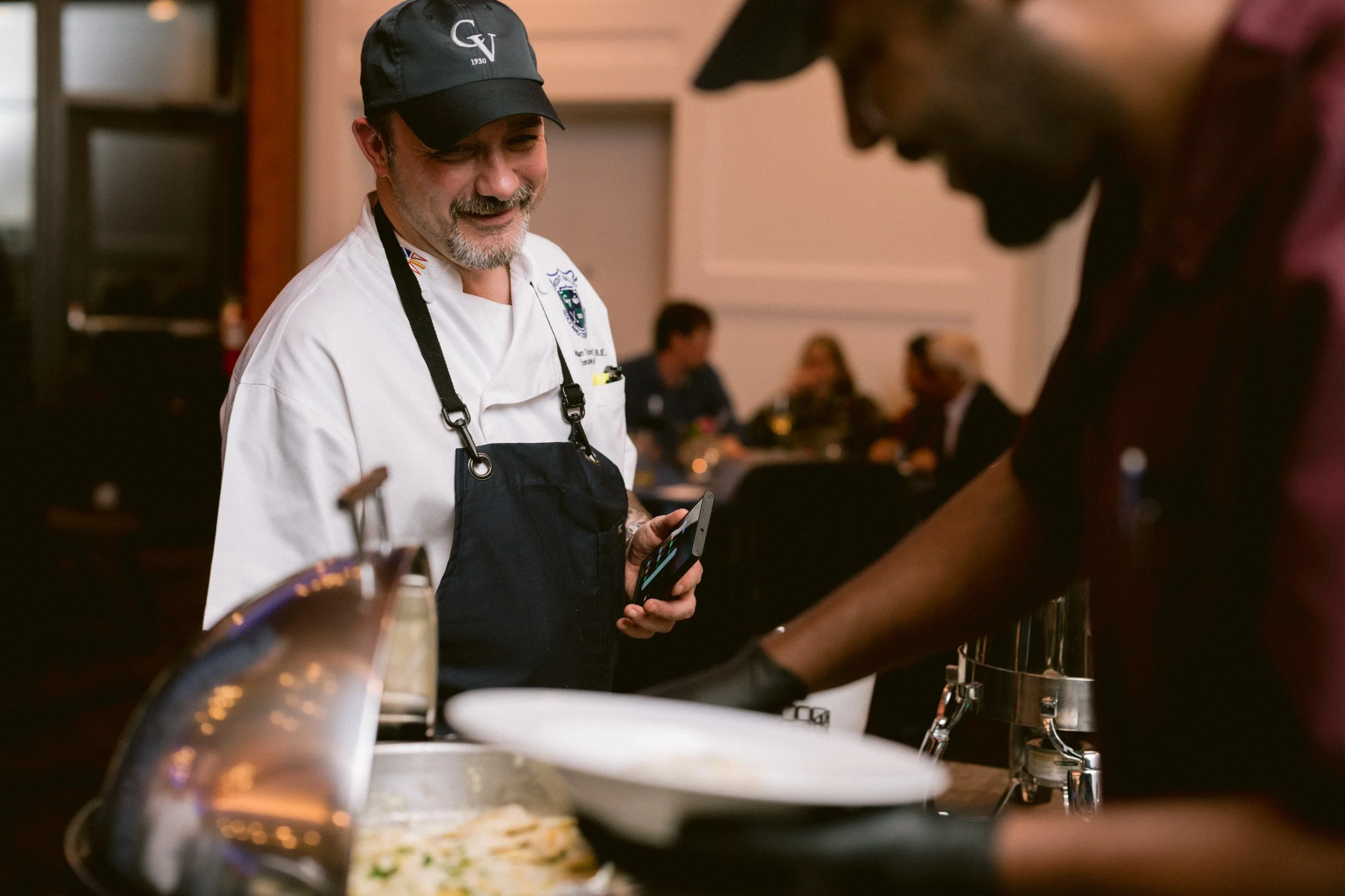 A smiling chef in a white uniform and black cap, holding a smartphone, at a restaurant serving food to a customer.
