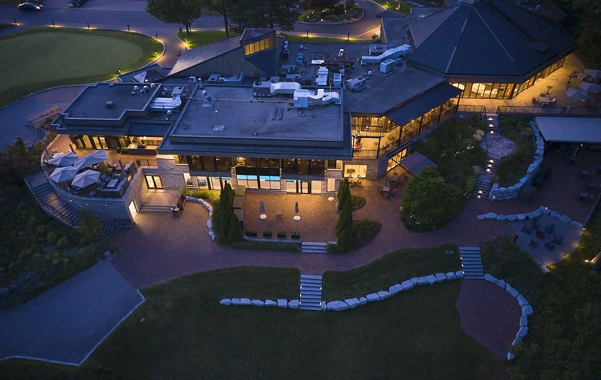 Aerial view of a modern house with outdoor lighting at dusk, showing a spacious patio, landscaped yard, and surrounding greenery.