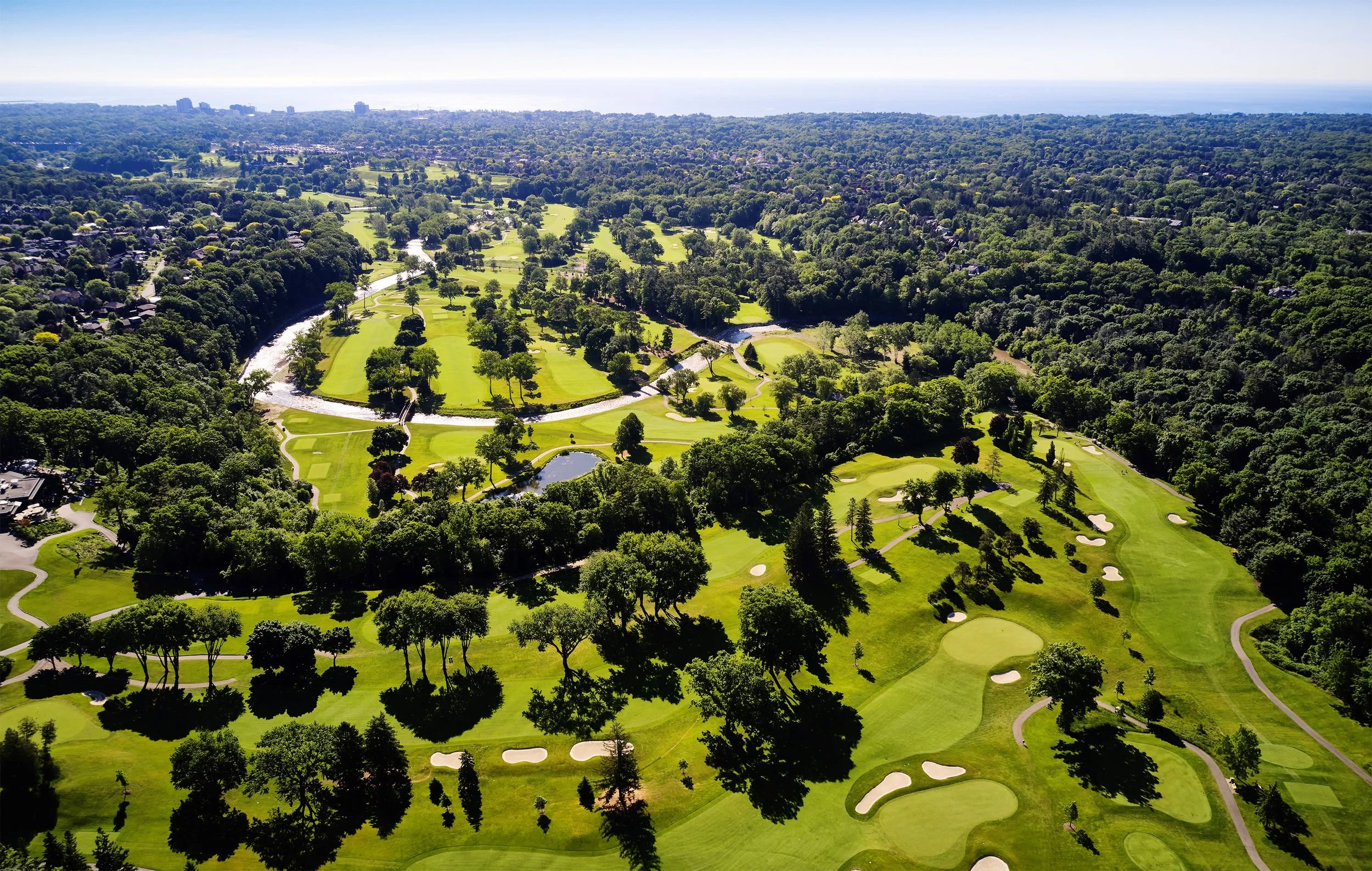 Aerial view of a lush golf course surrounded by dense trees, with small ponds and sand traps, extending toward Lake Ontario.