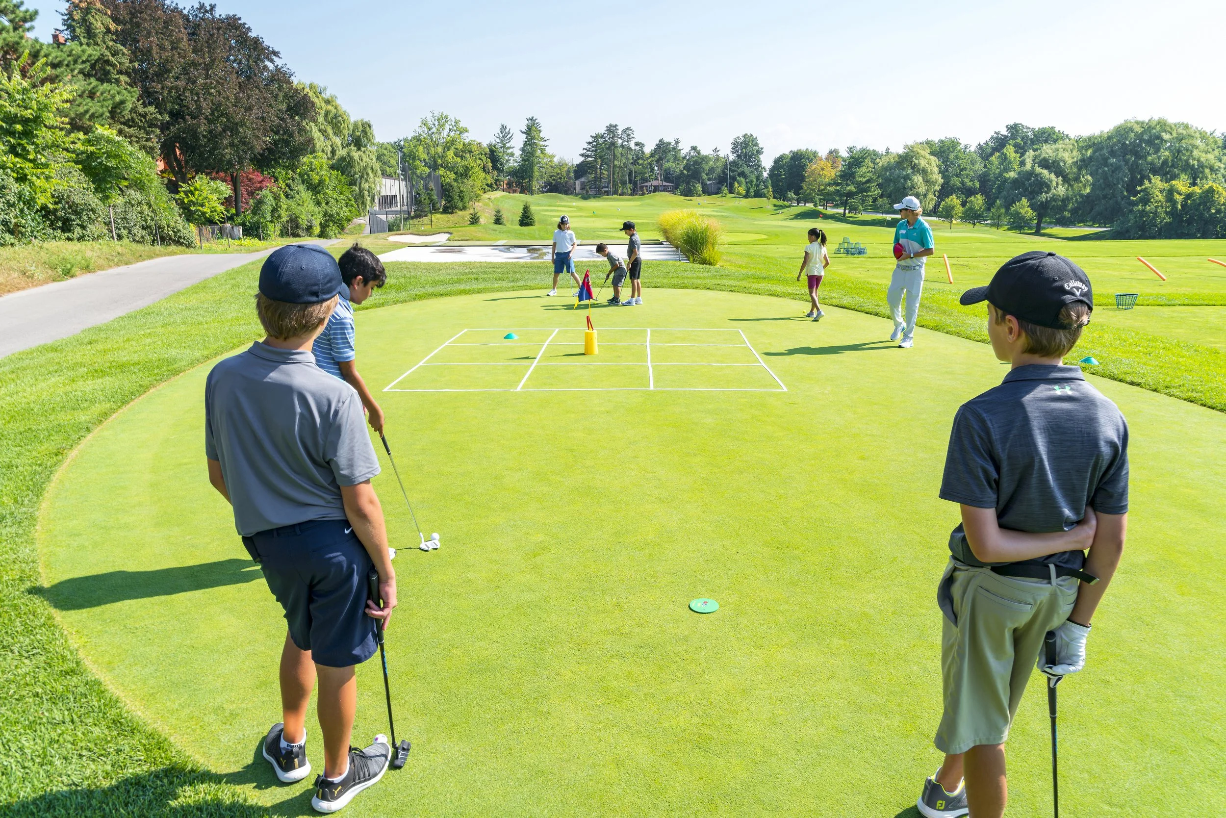 Children and adults playing golf on a sunny day at a golf course, with green grass, trees, and a clear blue sky.