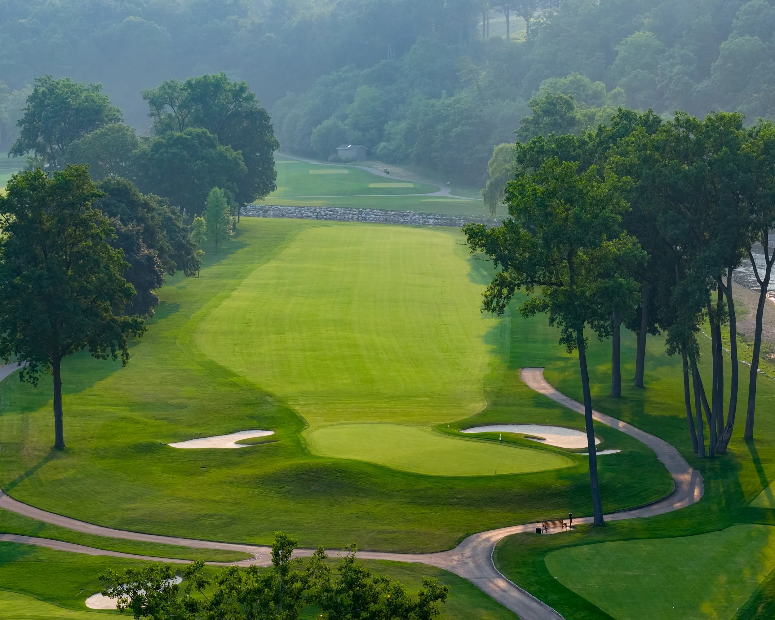 A scenic golf course with well-maintained green grass, sand bunkers, a paved cart path, and tall trees surrounding the area.