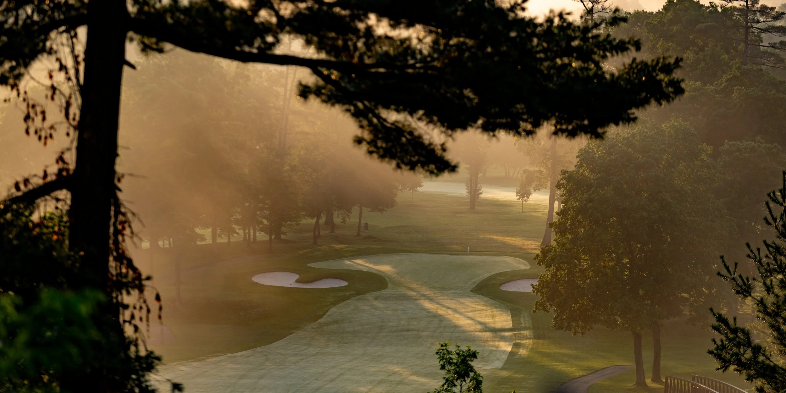 A golf course in the early morning with mist and sunlight filtering through trees.