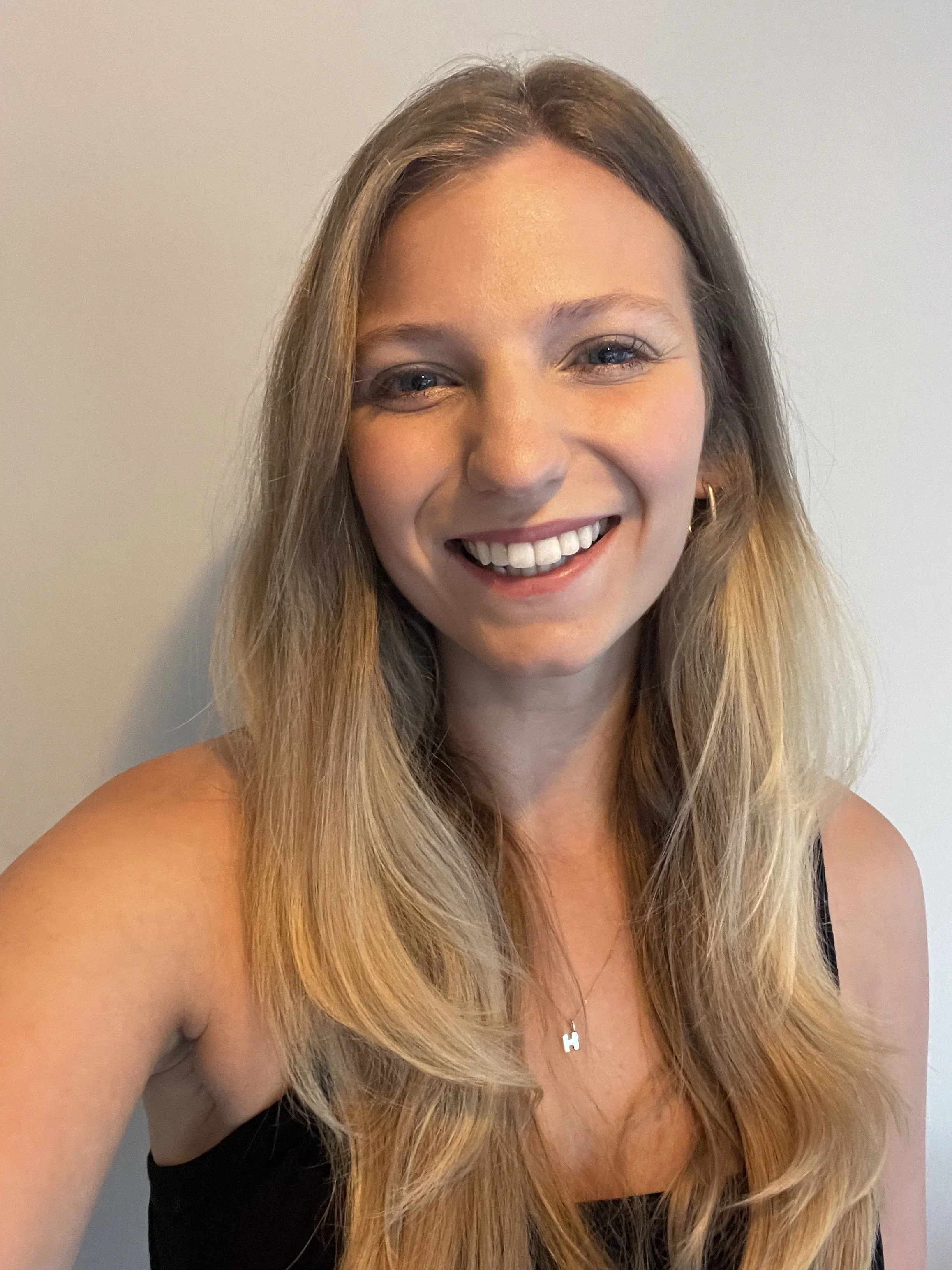 A young woman with long blonde hair, smiling, wearing a black top, a gold hoop earring, and a necklace with an 'H' pendant, standing against a plain light-colored wall.