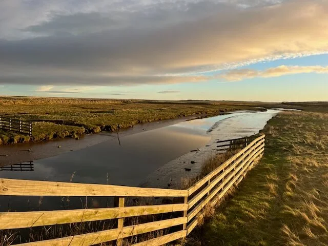Scenic rural landscape with a meandering river, wooden fences, grassy fields, and a dramatic cloudy sky.
