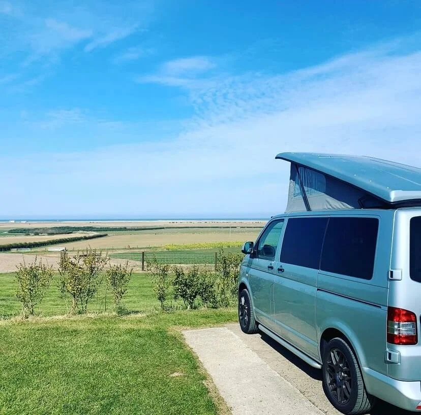 Campervan with pop-up roof parked on grassy area overlooking countryside and coast.