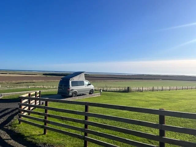 A camper van with a raised roof is parked on a green field near a wooden fence, overlooking a vast landscape under a clear blue sky.
