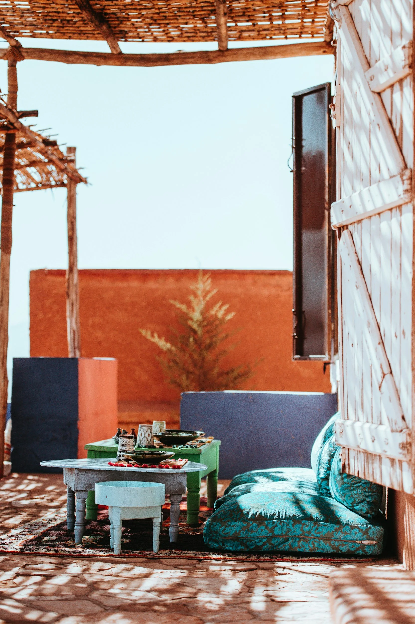 Outdoor seating area with colorful tables, cushions, and pottery under a wooden pergola, against a rustic wall backdrop.