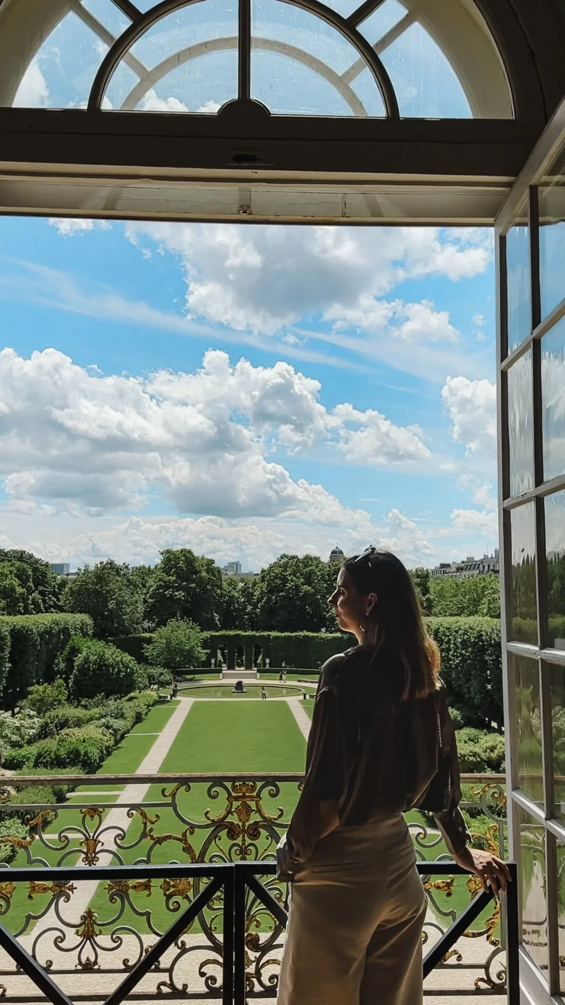 Woman standing on a balcony overlooking a garden with blue sky and clouds.