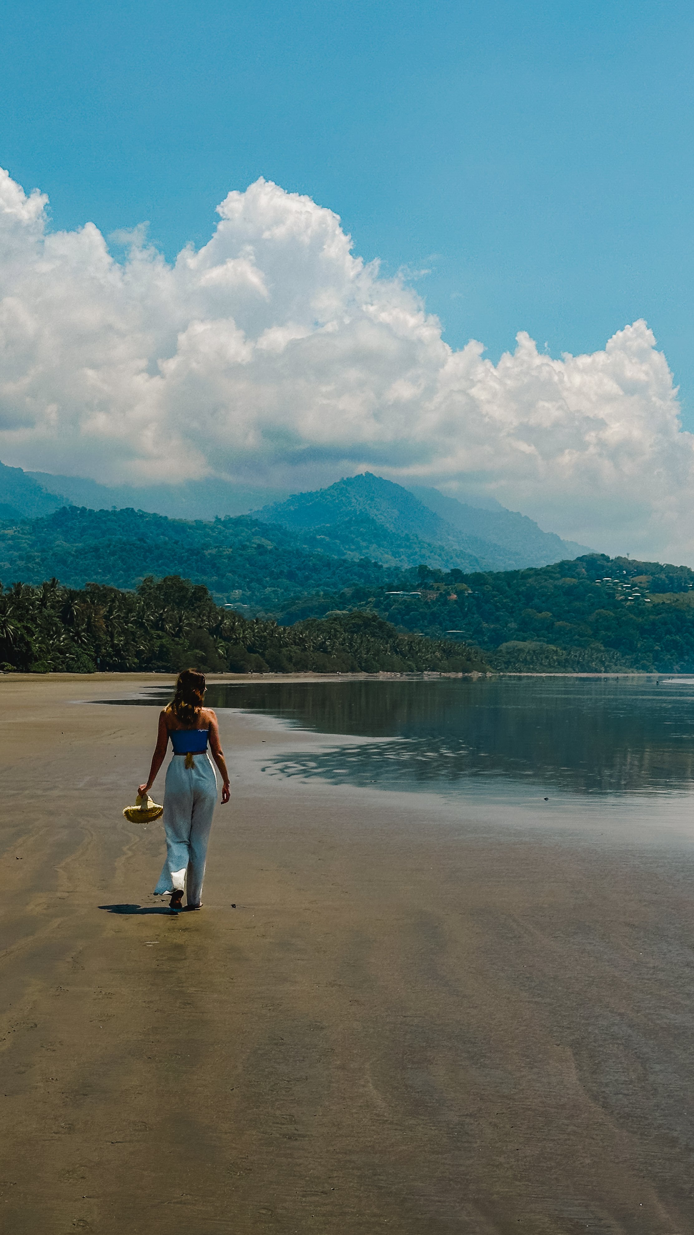 Person in swimwear standing on a beach with clear blue water and a mountain backdrop under a clear sky.