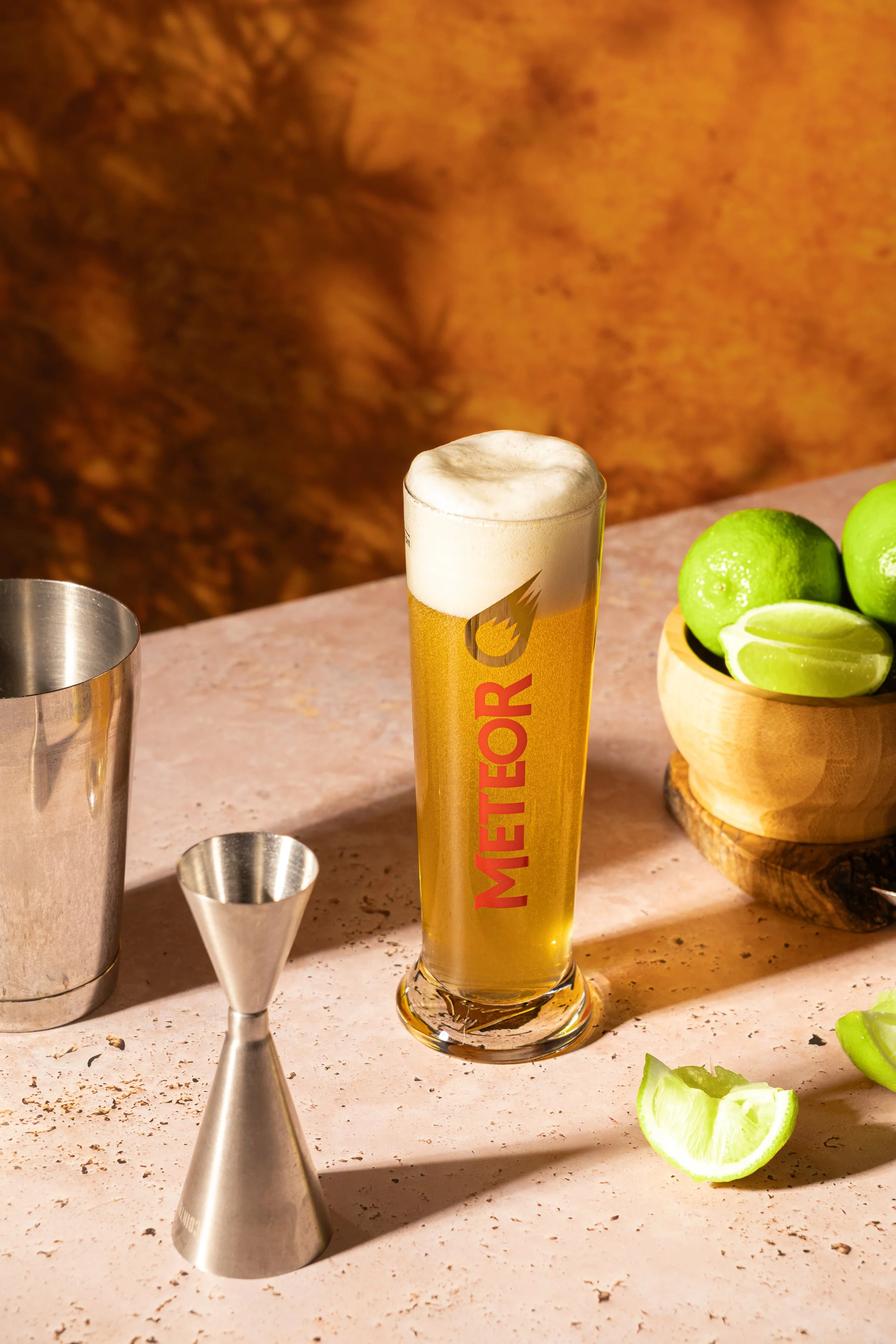 Verre de bière léger avec une mousse épaisse, citrons verts sur une planche en bois, et éléments de bar métalliques sur une surface en pierre, fond beige