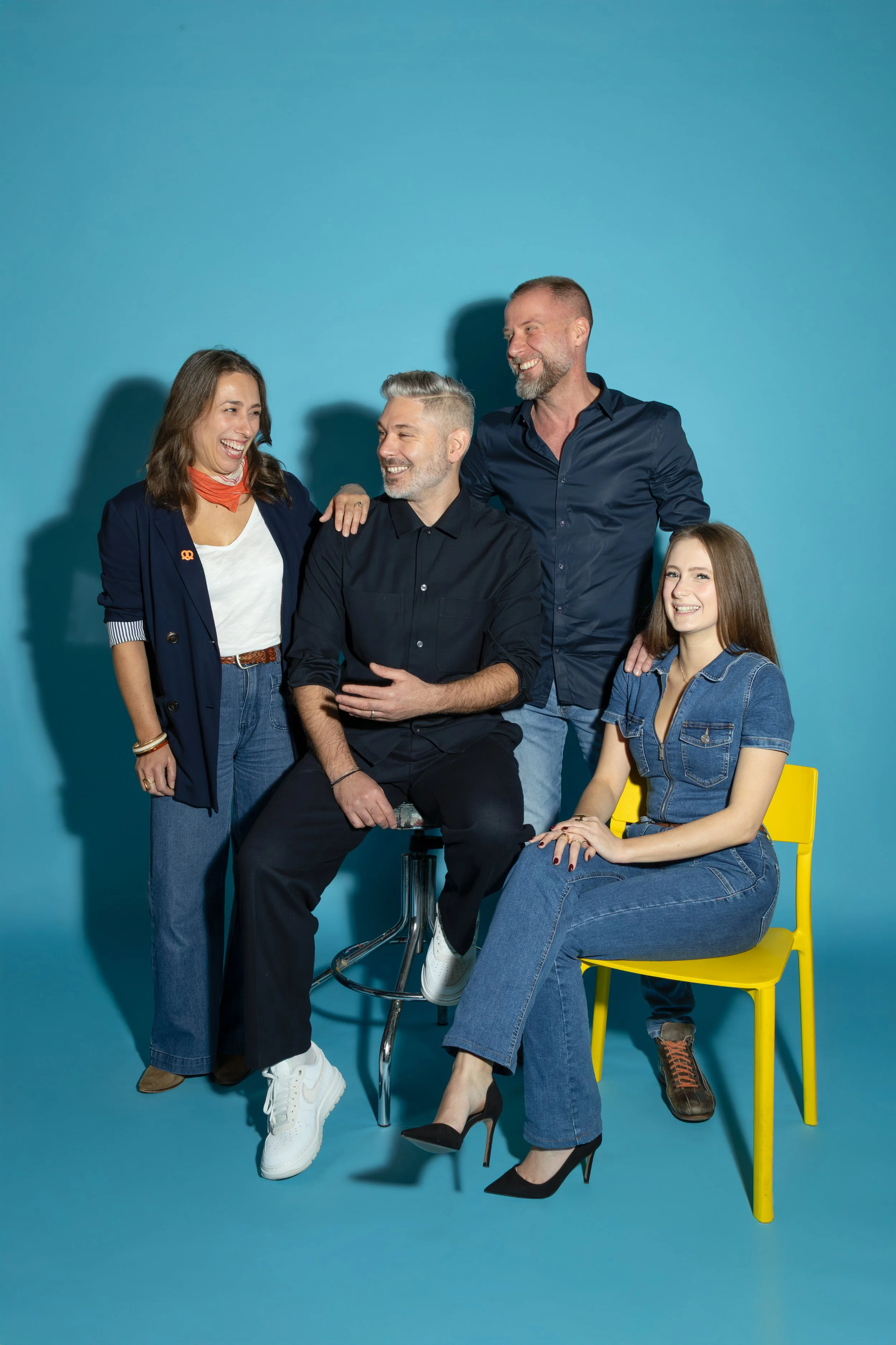 Cinq personnes souriantes interagissent dans une pièce avec un mur bleu, dont deux femmes et trois hommes, portant des vêtements décontractés et assises ou debout près d'une chaise jaune.