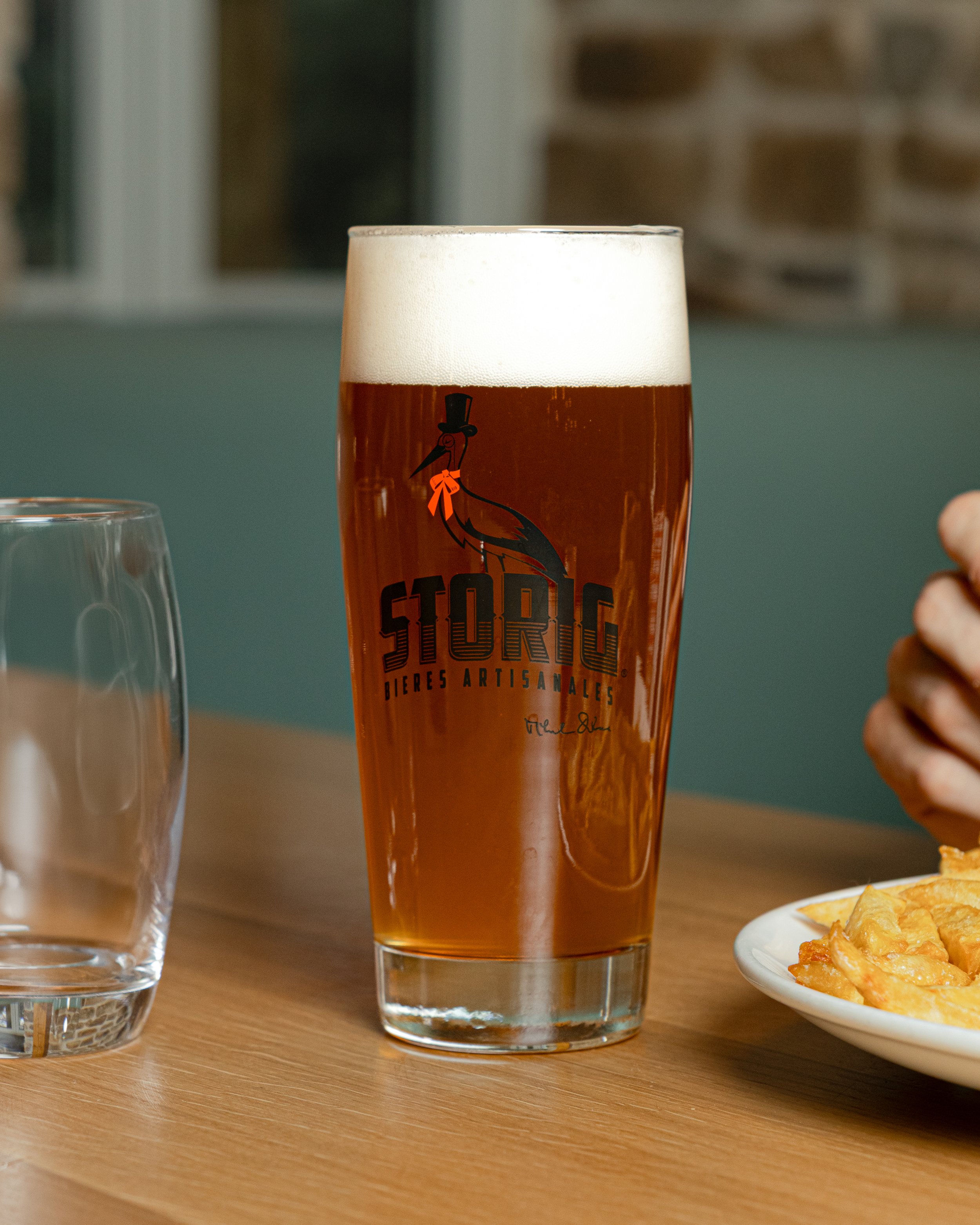 Verre de bière artisanale avec un logo de cigogne portant un chapeau haut de forme et une cravate orange, placé sur une table en bois, avec un verre d'eau à gauche et une assiette de chips à droite.