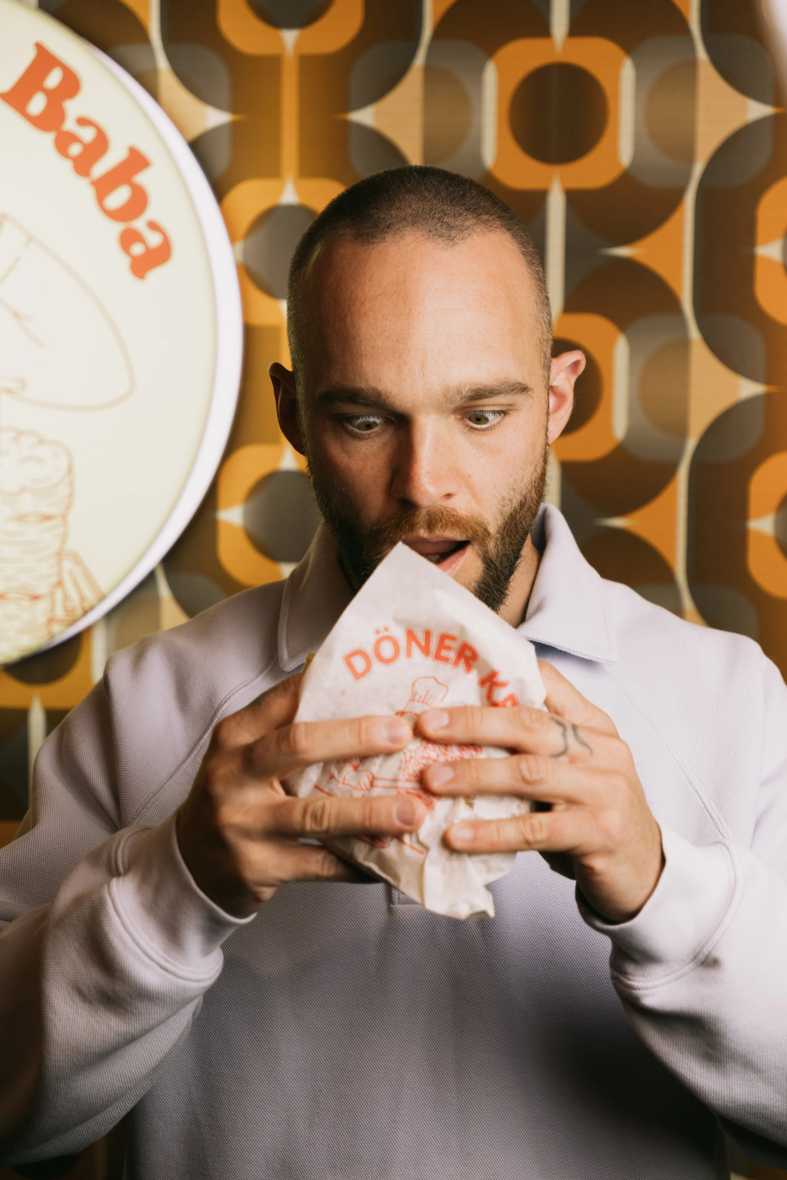 Un homme à la barbe et sans cheveux apparents tient un sandwich dans ses mains, l'air surpris ou impressionné. En arrière-plan, un motif géométrique coloré avec du orange, marron et beige, et un panneau circulaire partiel avec du texte en allemand.