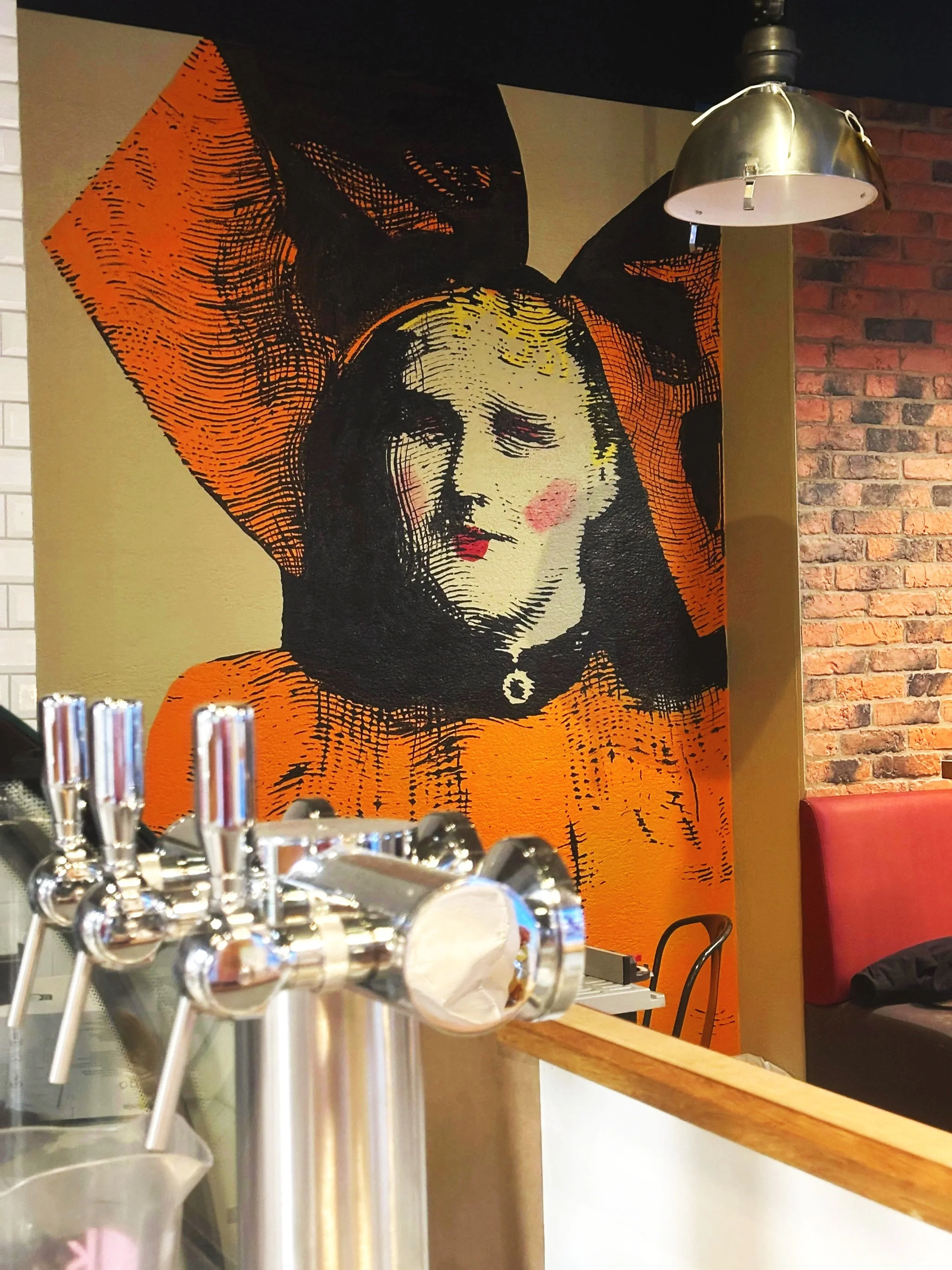Mur en briques avec une grande affiche de style pop art représentant une femme avec une coiffure volumineuse, portant un collier et un vêtement orange.