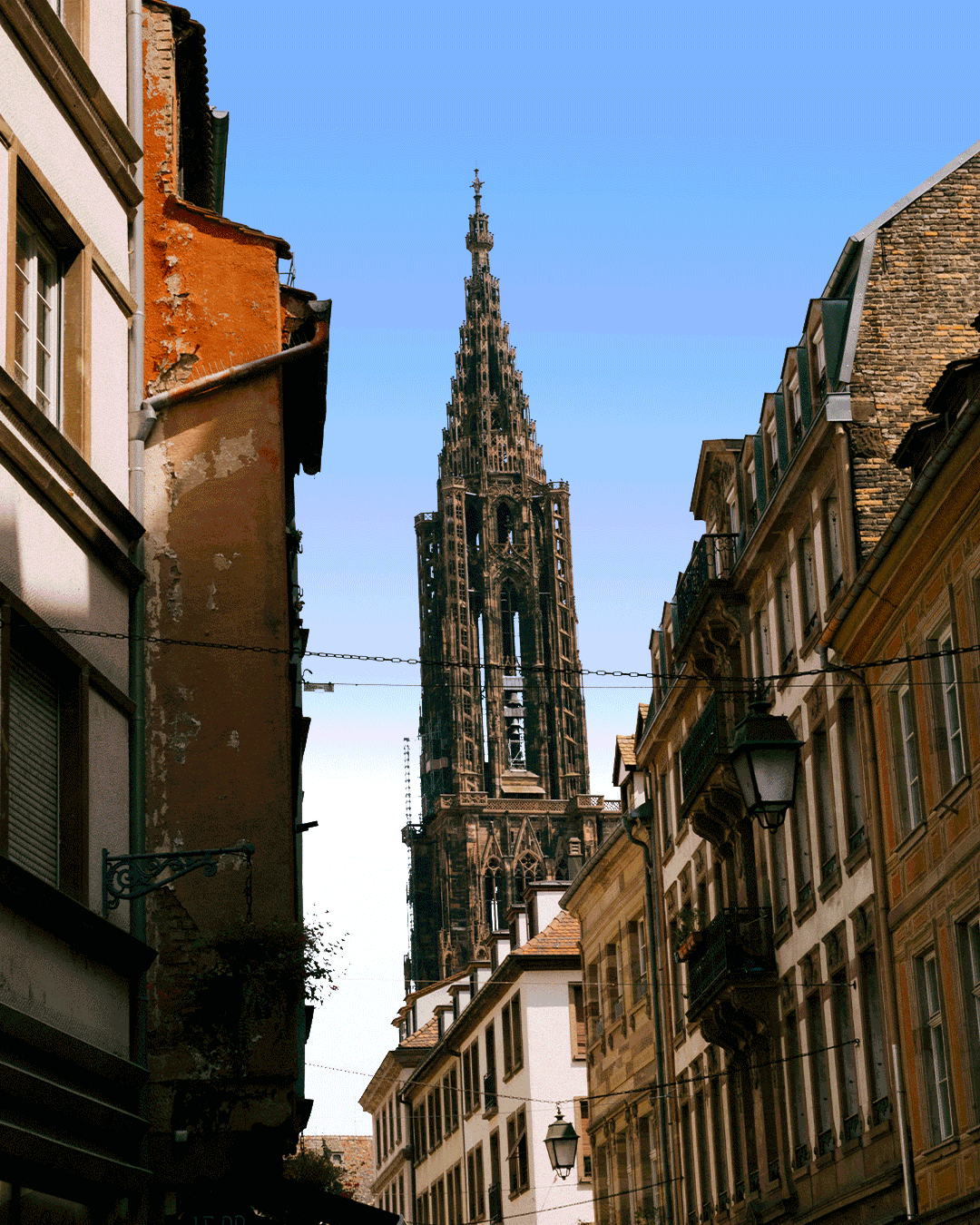 Vue d'une rue étroite avec des bâtiments anciens et la cathédrale de Strasbourg en arrière-plan, sous un ciel bleu.