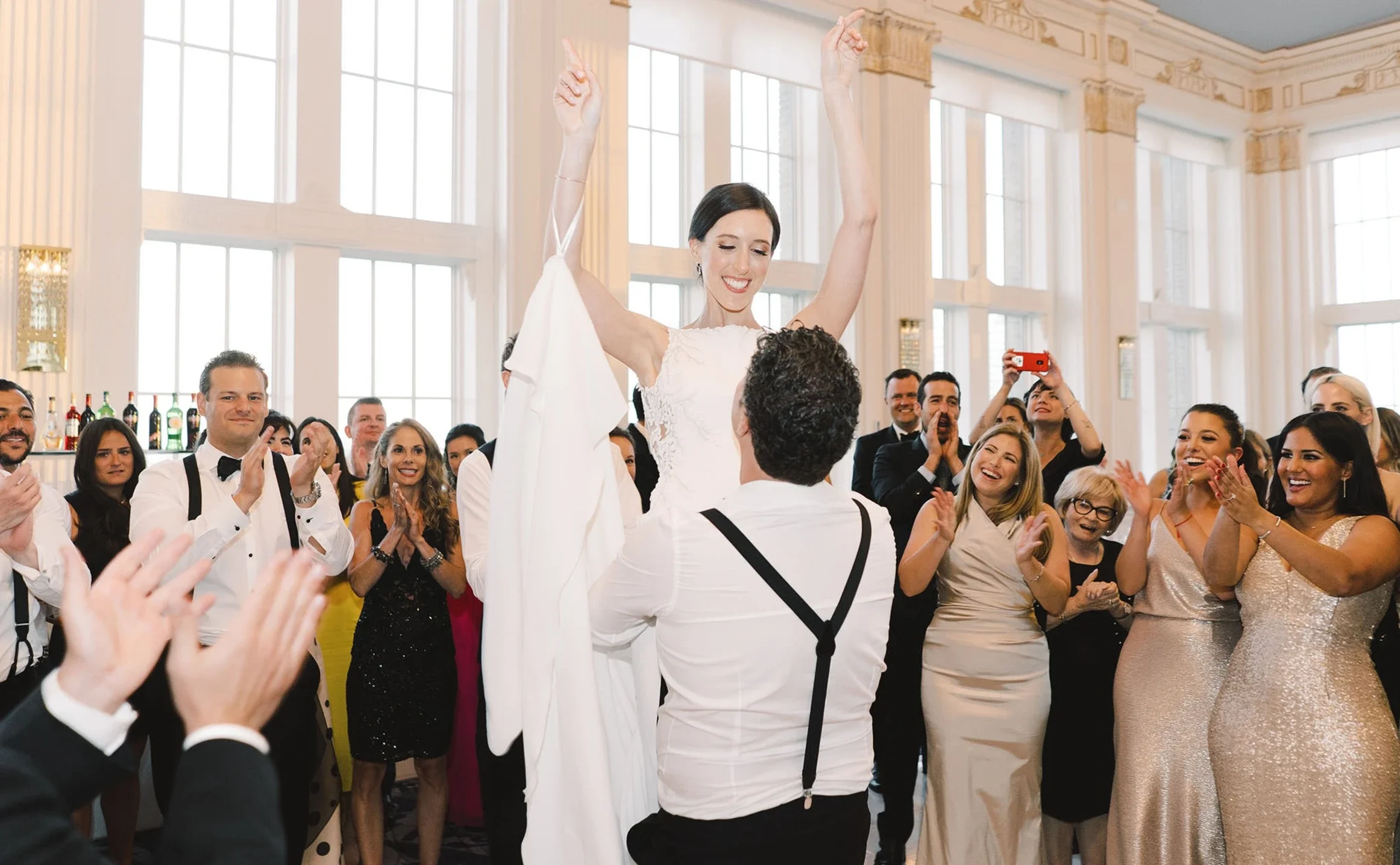 Group of people dancing and celebrating at a wedding reception, with some holding drinks and others with arms raised, inside a decorated tent.