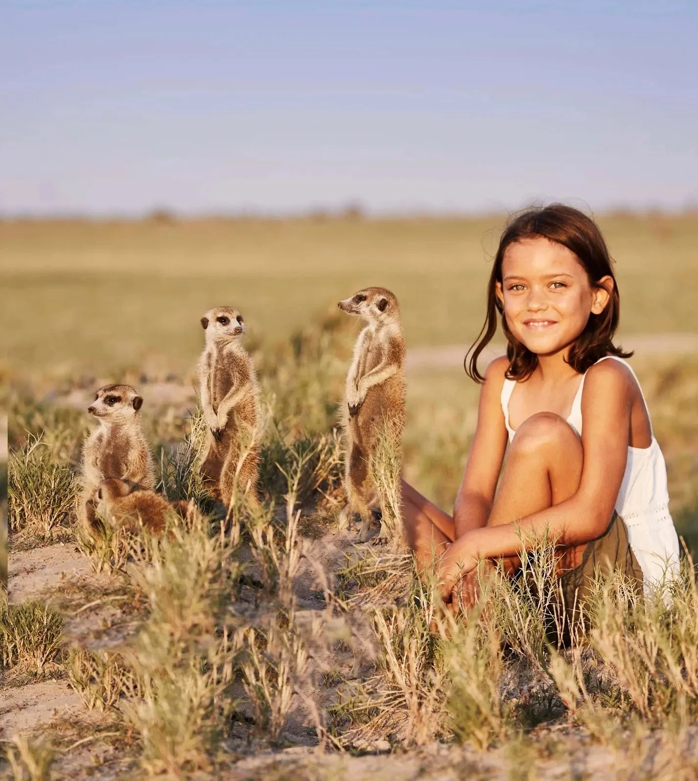 Young girl sitting in grass next to three meerkats standing upright in a field.