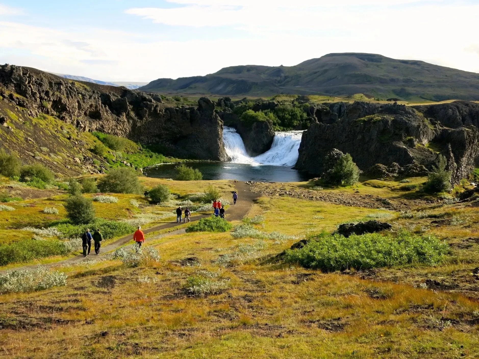 Scenic landscape with a waterfall surrounded by rocky cliffs and lush greenery, people walking on a path in the foreground, mountains in the background.