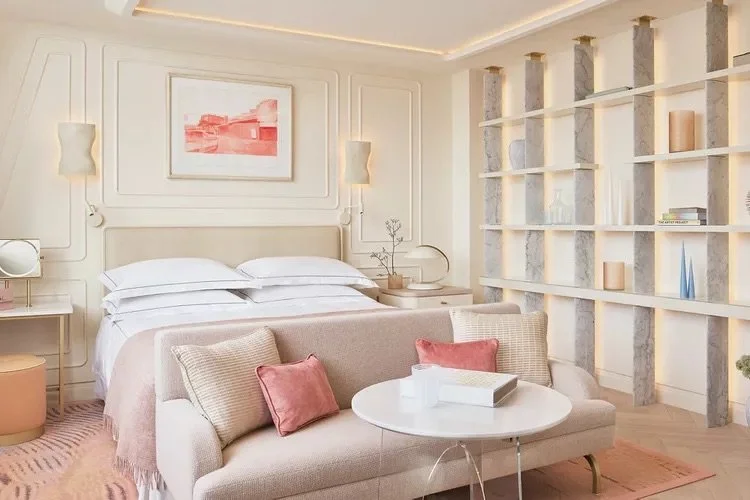 Luxurious hotel room with a beige and pink color scheme, featuring a bed with white linens, a stylish sofa, and a round coffee table. The wall has an abstract art piece, and there's a decorative shelving unit on the side.