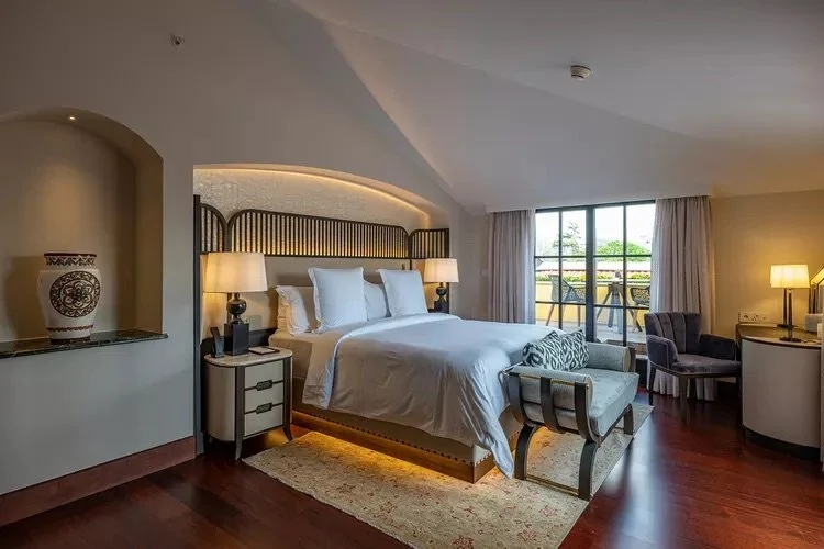 Luxury hotel room featuring a large bed with white linens, accent lamps, a decorative vase on a shelf, and a sitting area with chairs beside a window overlooking a balcony.
