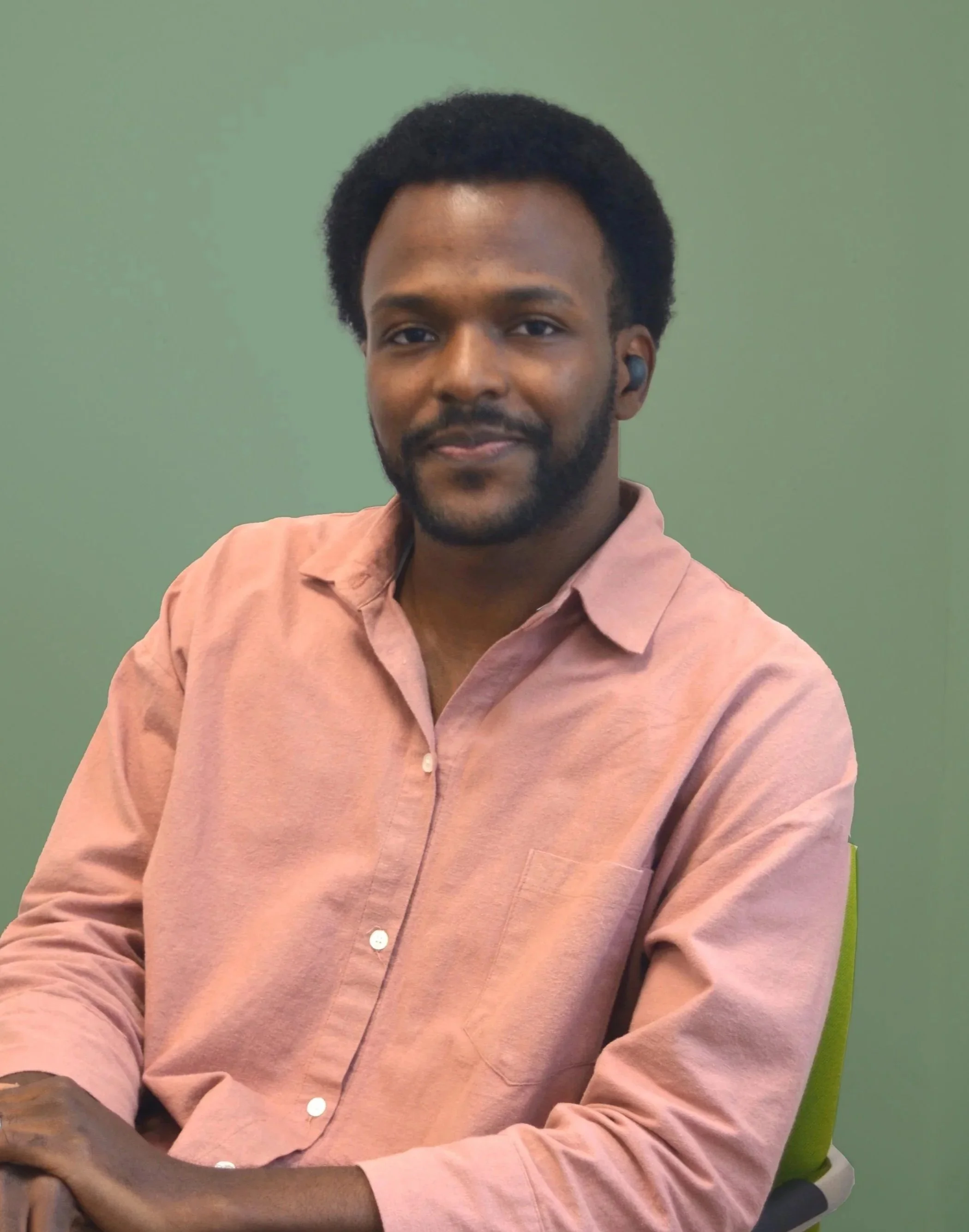 A young man with dark, curly hair and a beard sitting on a green chair against a green background, wearing a pink button-up shirt with arms crossed.