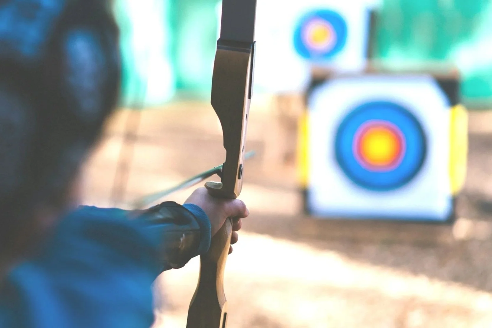 A person aiming a bow with an arrow towards a target in an indoor archery range.
