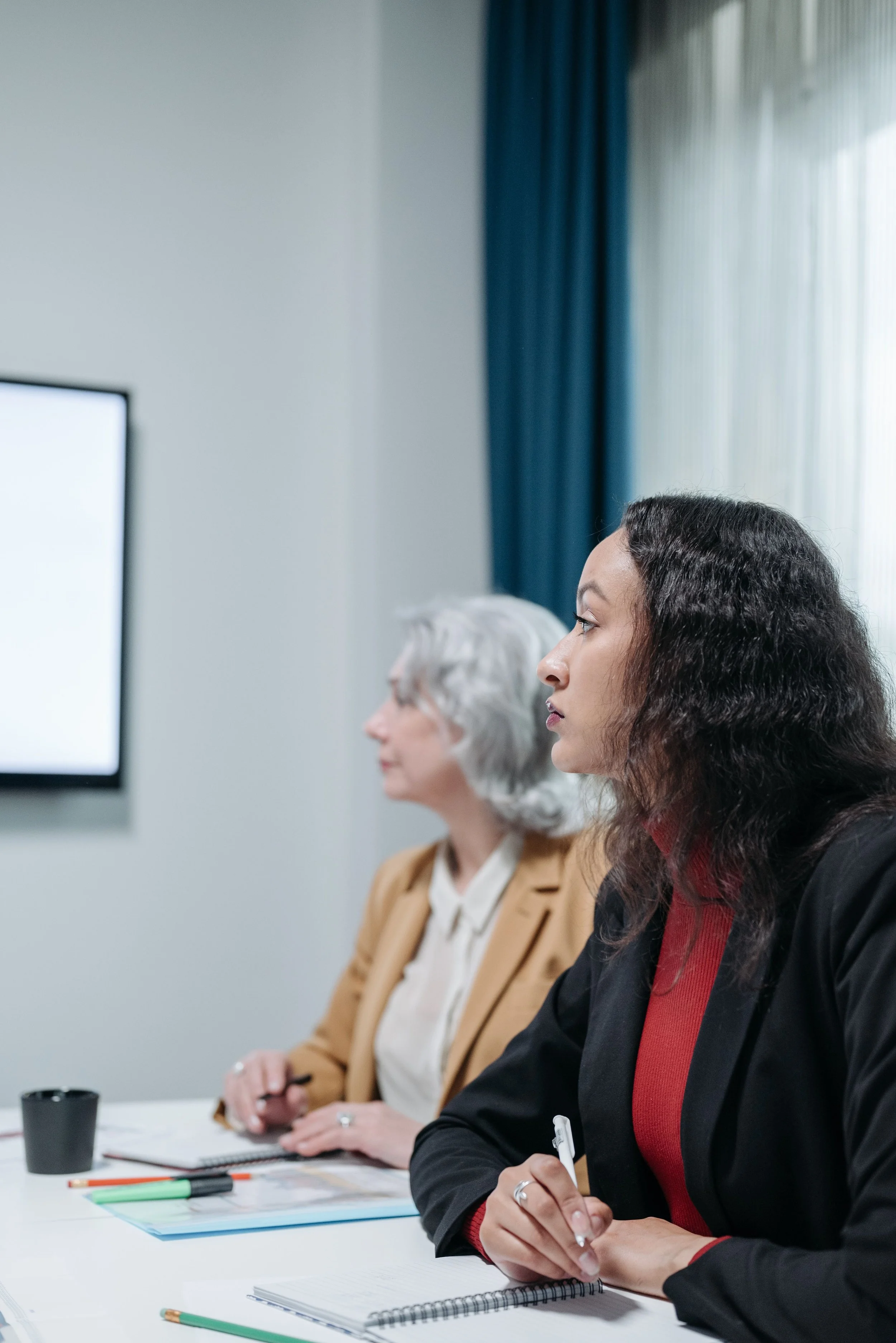 Three women in business attire attending a meeting or conference, sitting at a white table with notebooks, pens, and a cup on it. They are looking at a screen or presentation off-camera.