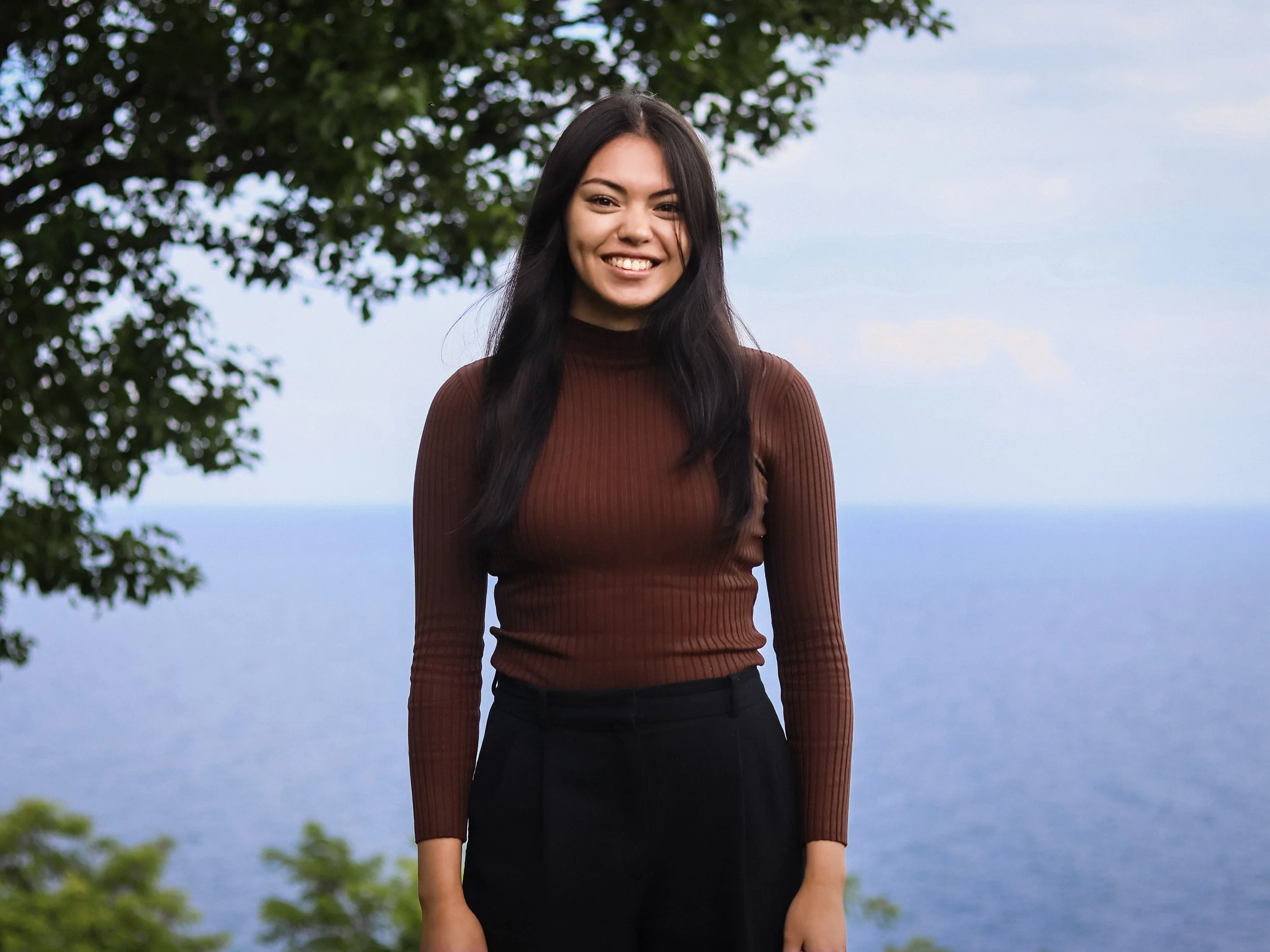 A young woman with long dark hair stands outdoors near a body of water, smiling at the camera with trees in the background.