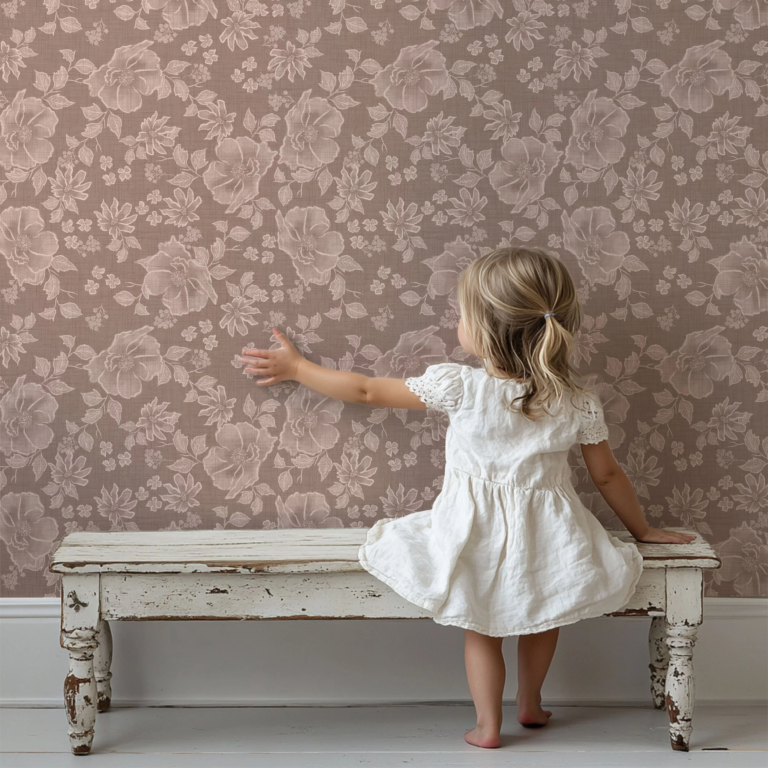 A young child in a white dress stands barefoot on a rustic wooden bench, reaching toward a dusty rose floral wallpaper with delicate cream botanical patterns. The soft neutral tones and vintage-inspired design create a calm, nostalgic atmosphere.