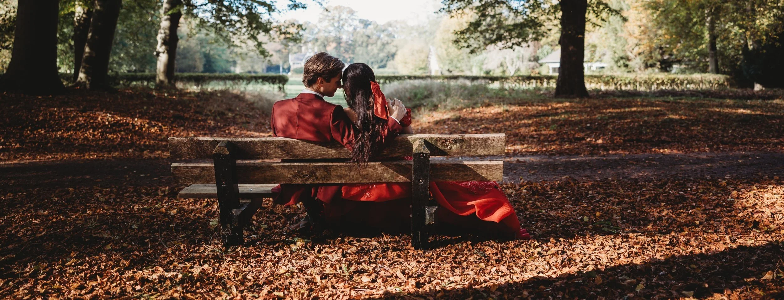 Bruidspaar in rode kleding zitten op een bank in een park met herfstbladeren, omringd door bomen met groen en herfstkleuren op hun bruiloft