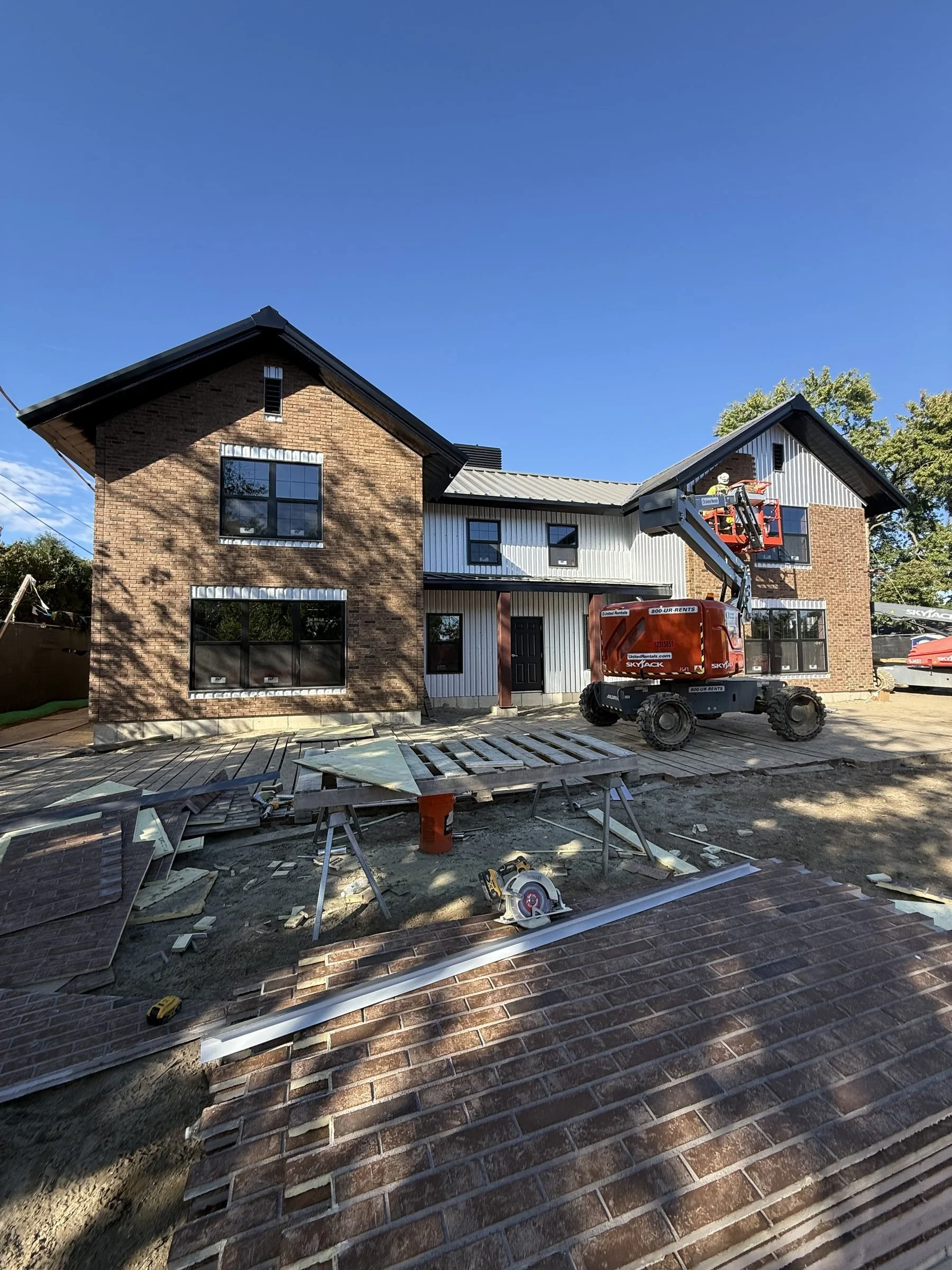 Brick building under construction with a boom lift, construction materials, and partially completed brick paving under a clear blue sky.