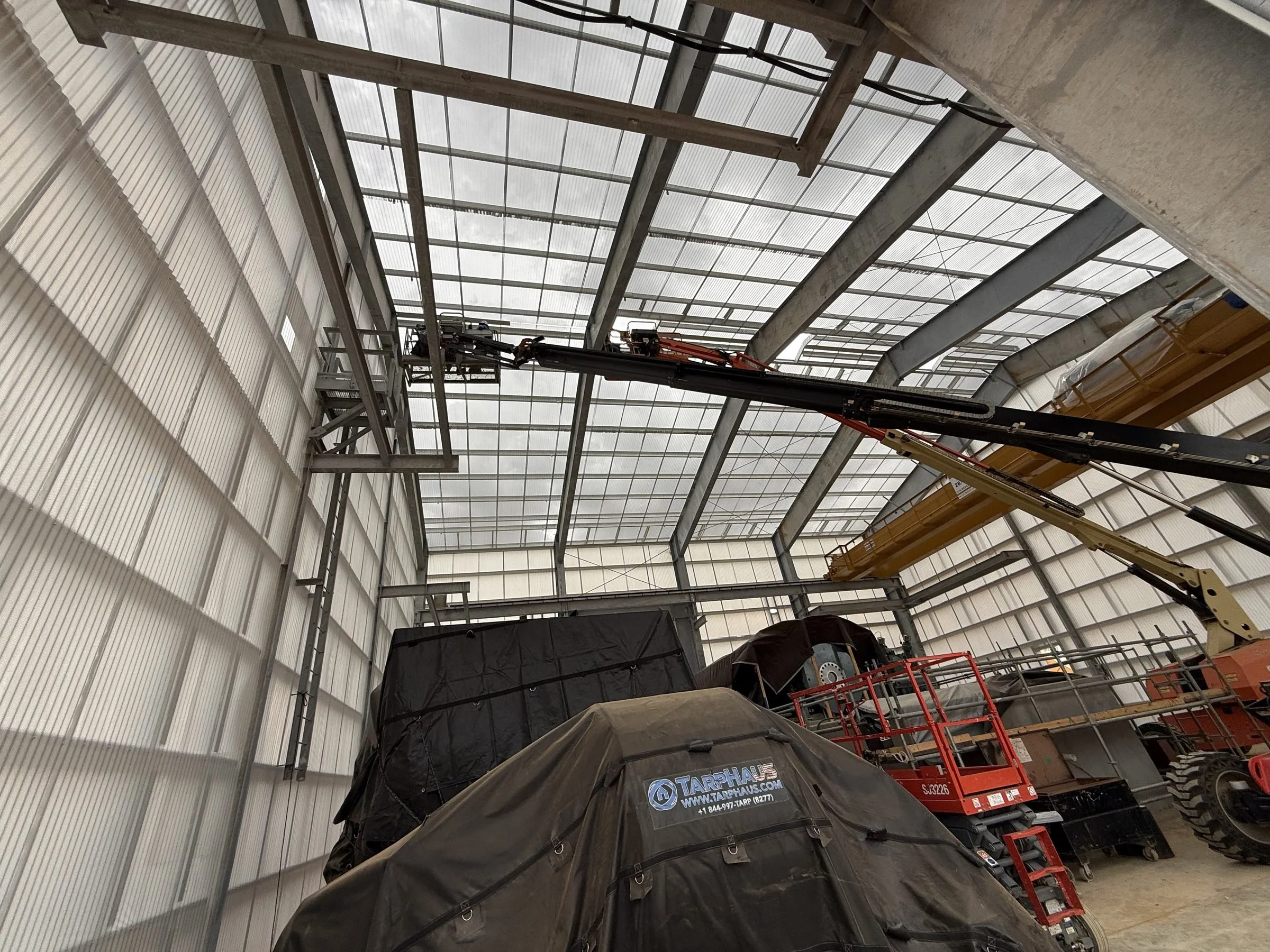 Industrial workshop interior with heavy machinery, covered equipment, aerial work platforms, and a translucent roof supported by steel beams