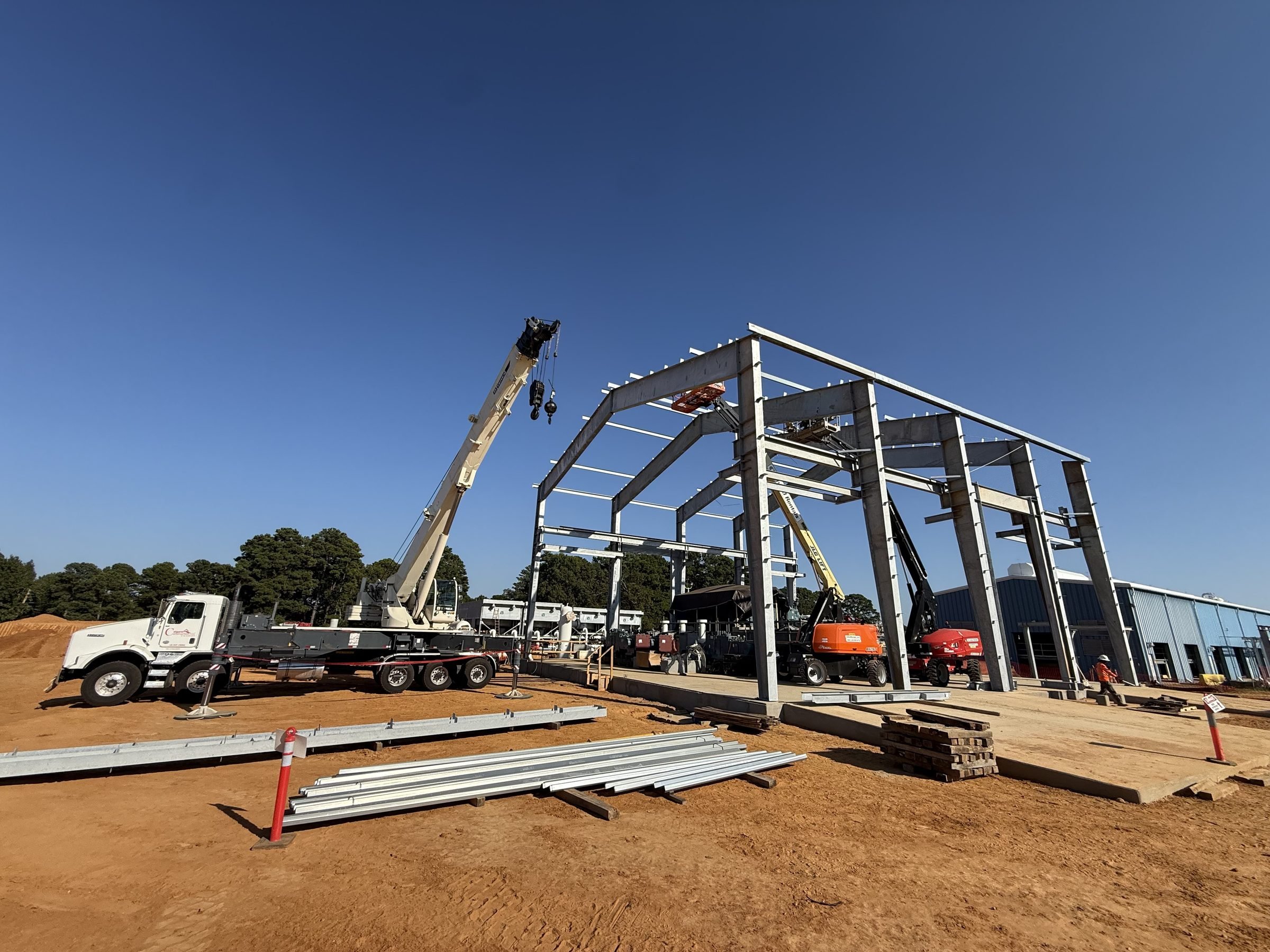 Steel-frame commercial building under construction with cranes, excavators, construction materials, and workers on a sunny day
