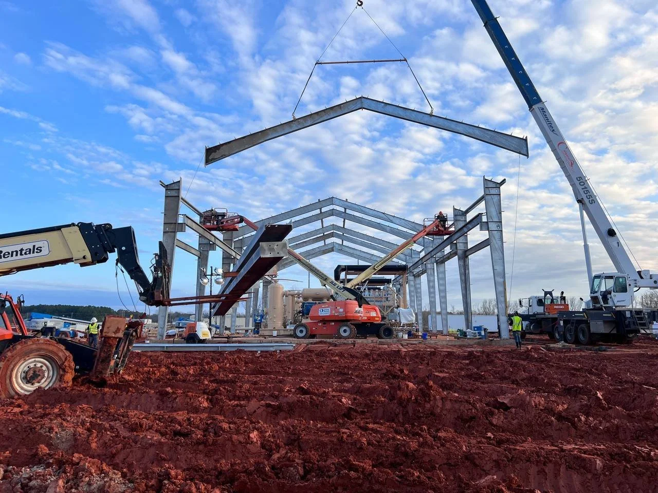 Steel building construction site with a crane lifting a roof beam, aerial lifts, PPE compliant workers.