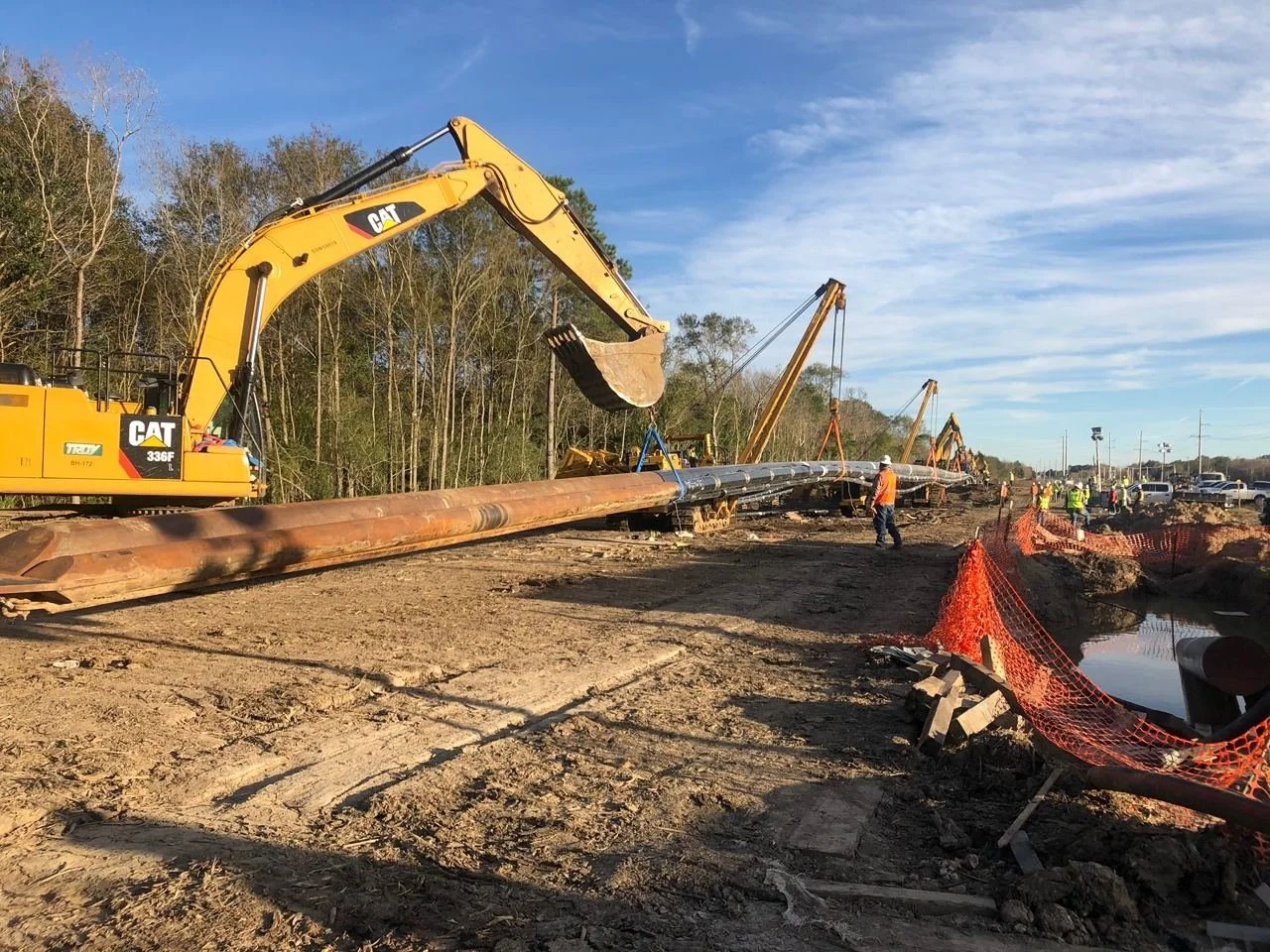 CAT excavator lifting a large pipeline at a roadside construction site with workers and cranes.