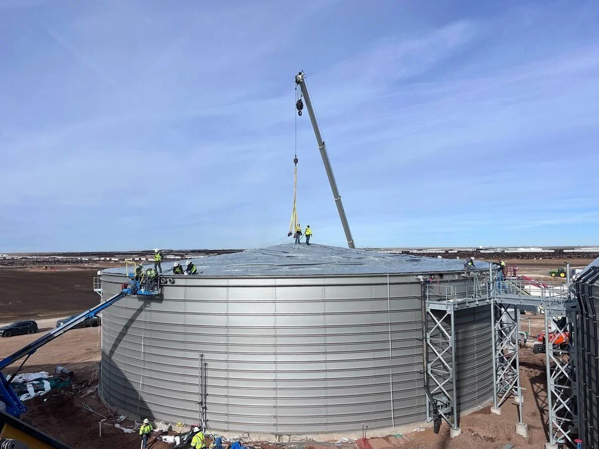 Construction workers install a domed roof on a large industrial biogas tank using a crane.