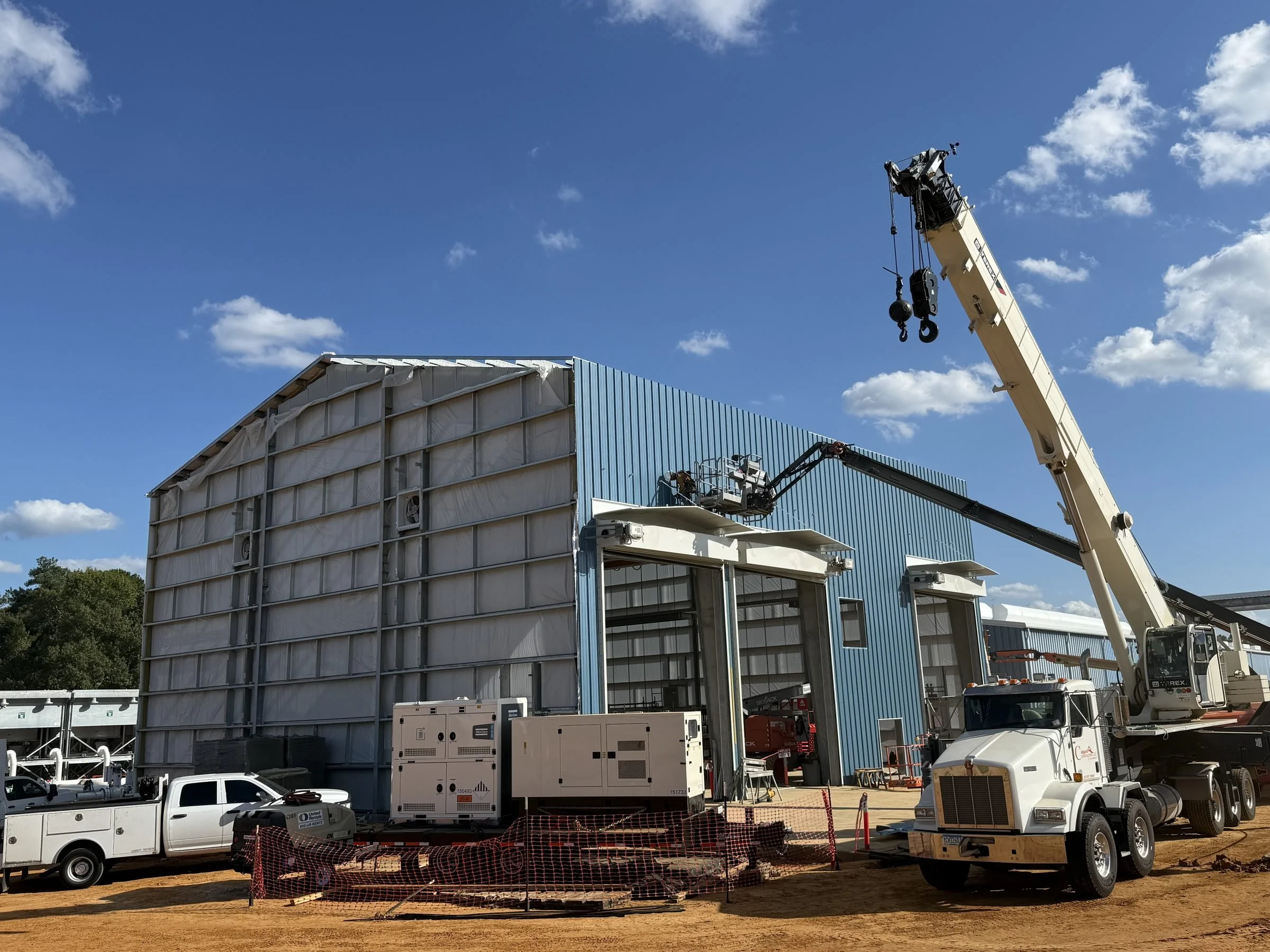 Construction site with a blue industrial building, mobile cranes, aerial lifts, generators, and construction vehicles under a clear blue sky.