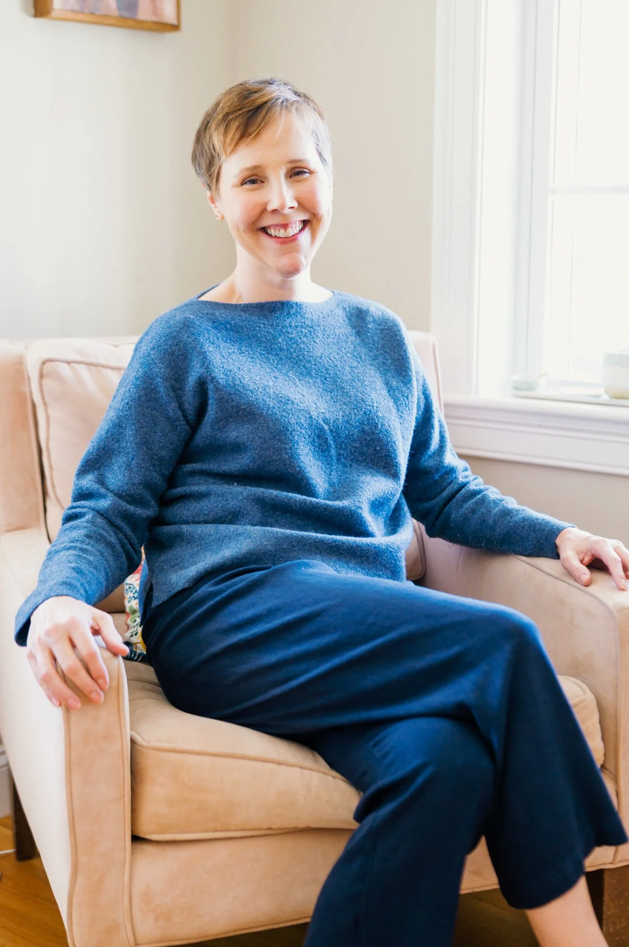 A woman with short reddish hair and a big smile, wearing a blue sweater and dark pants, sitting on a beige sofa in a bright room with white walls and a window.