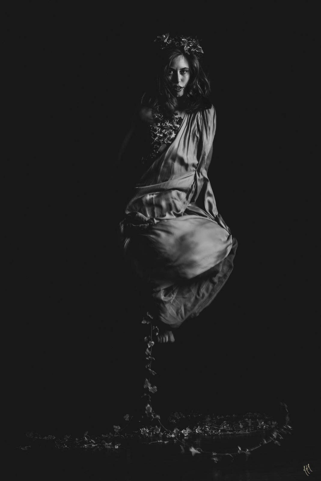 Black and white photo of a woman in a flowing dress and floral headpiece, appearing to float, surrounded by cascading vines.