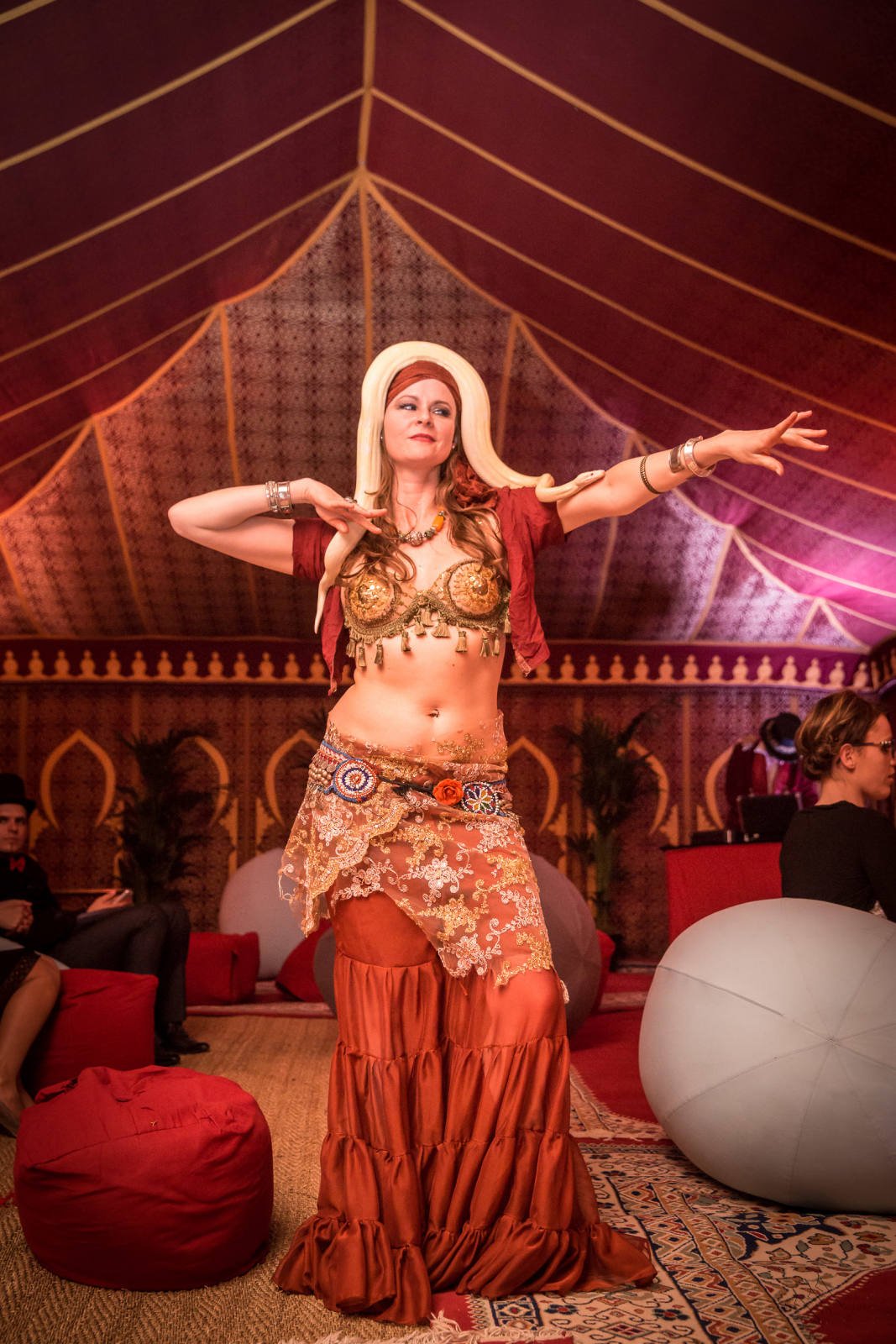 A woman in an ornate costume performing a dance inside a tent, wearing a headband and balancing a snake on her head and arms. The setting has a festive, decorated interior with people seated in the background.