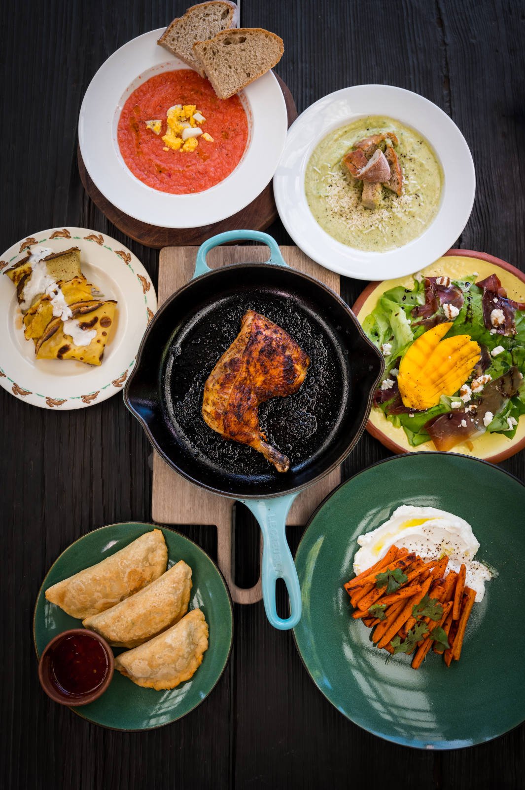 A variety of dishes on a dark wooden table, including a cast iron skillet with roasted chicken, a bowl of tomato soup with bread, a plate with three empanadas and dipping sauce, grilled squash, a salad with mango and cheese, a plate of seasoned carro