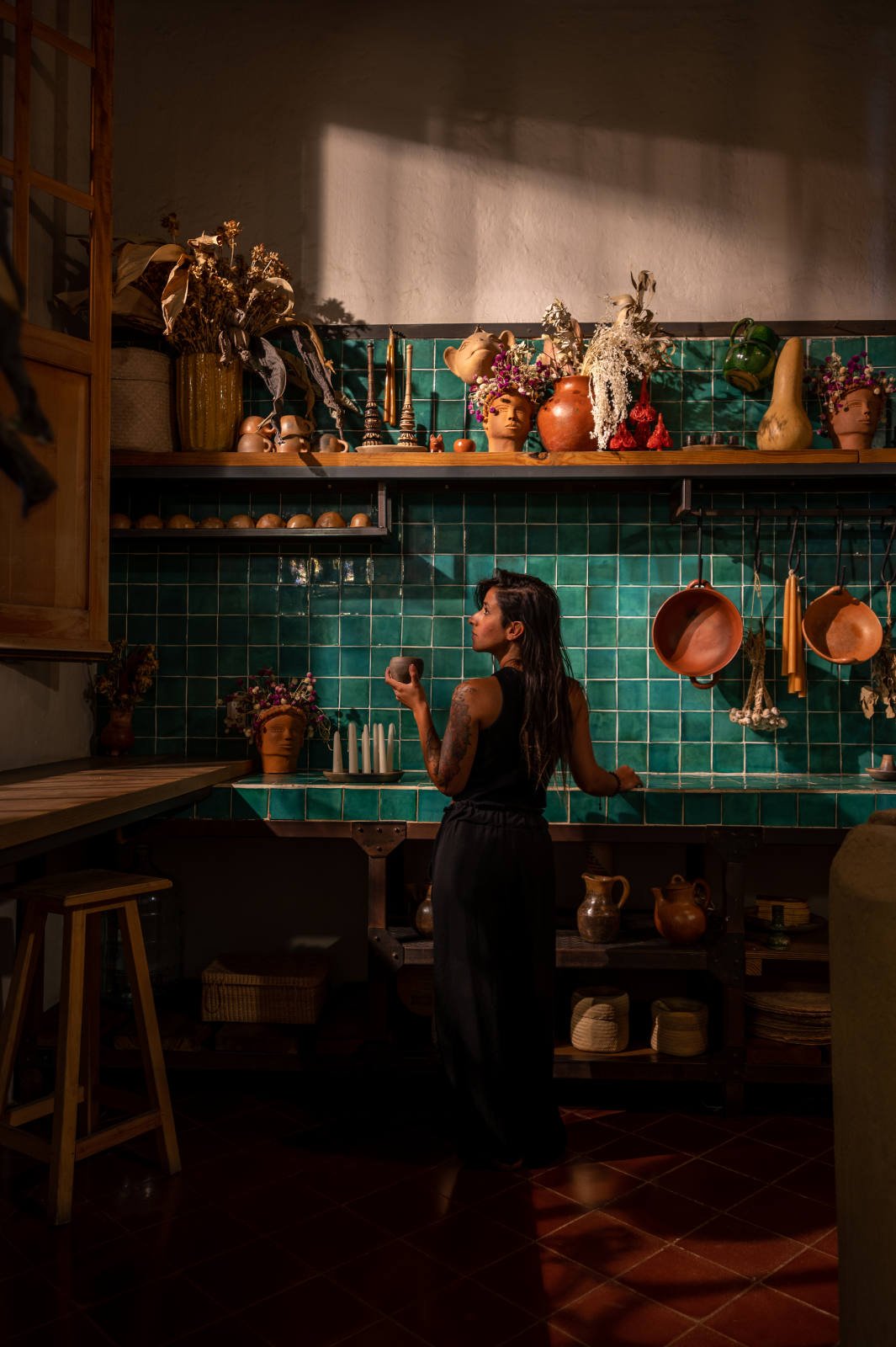 Woman holding a cup in a rustic kitchen with green tiled wall, shelves filled with decorative pottery and dried flowers, hanging pots and pans, and a wooden countertop.