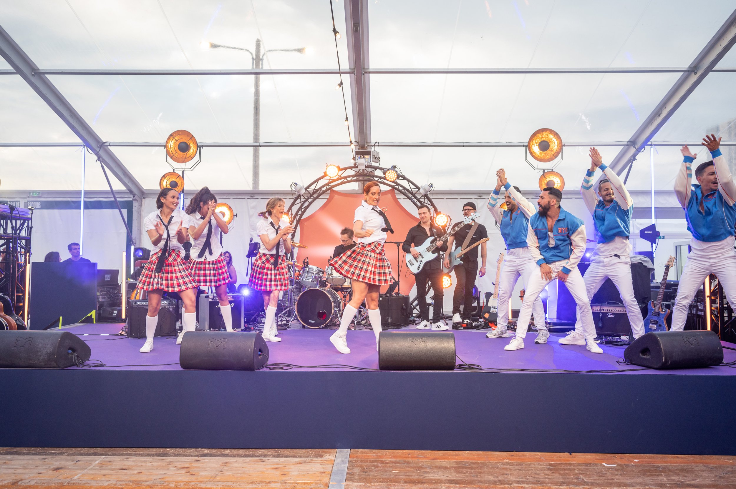 Performers dancing on stage under a transparent tent, wearing red plaid skirts and blue jackets. Musical instruments and stage lights are visible in the background.