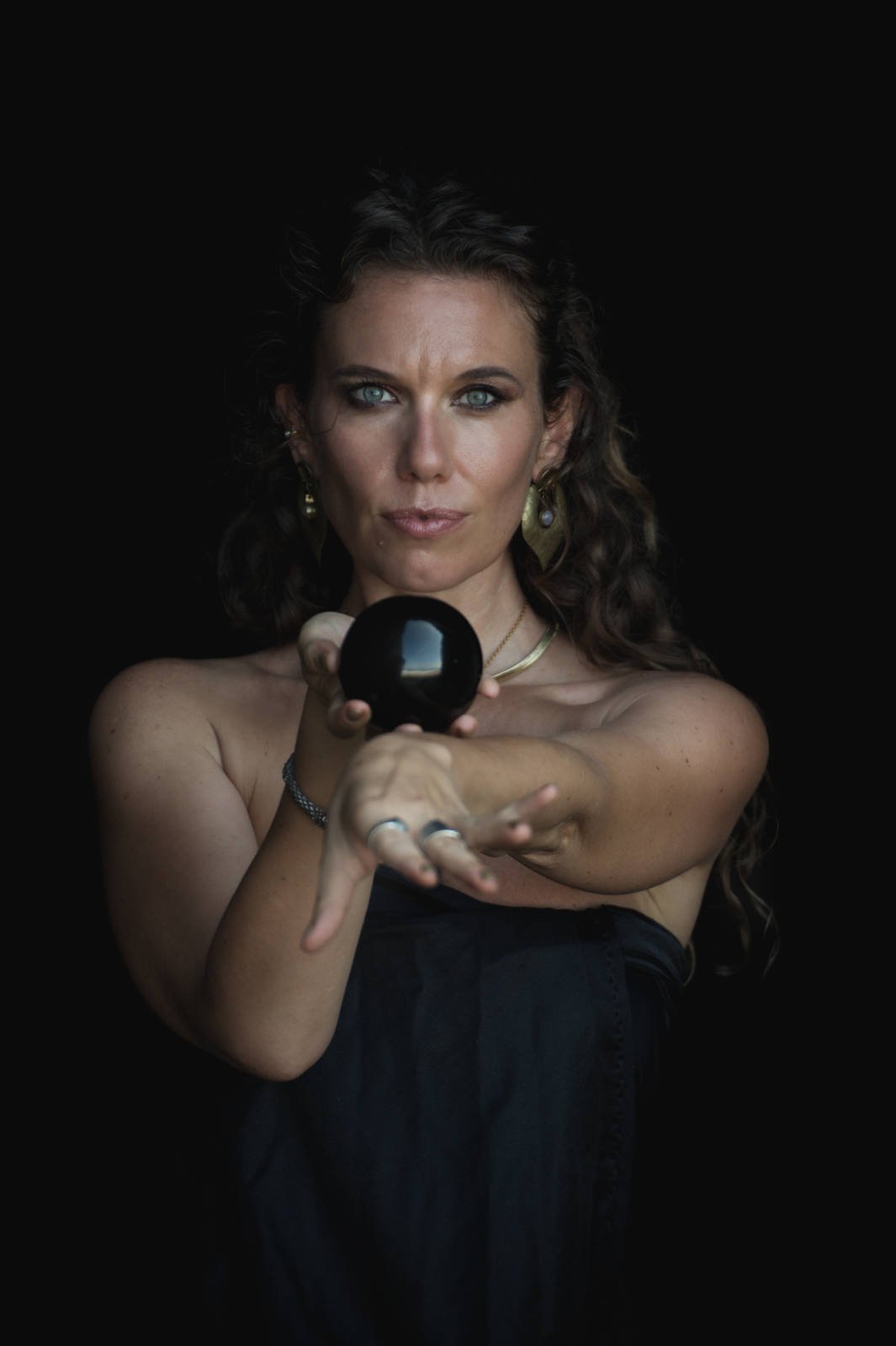 Woman holding a black sphere against a dark background, wearing earrings and jewelry, focused expression.